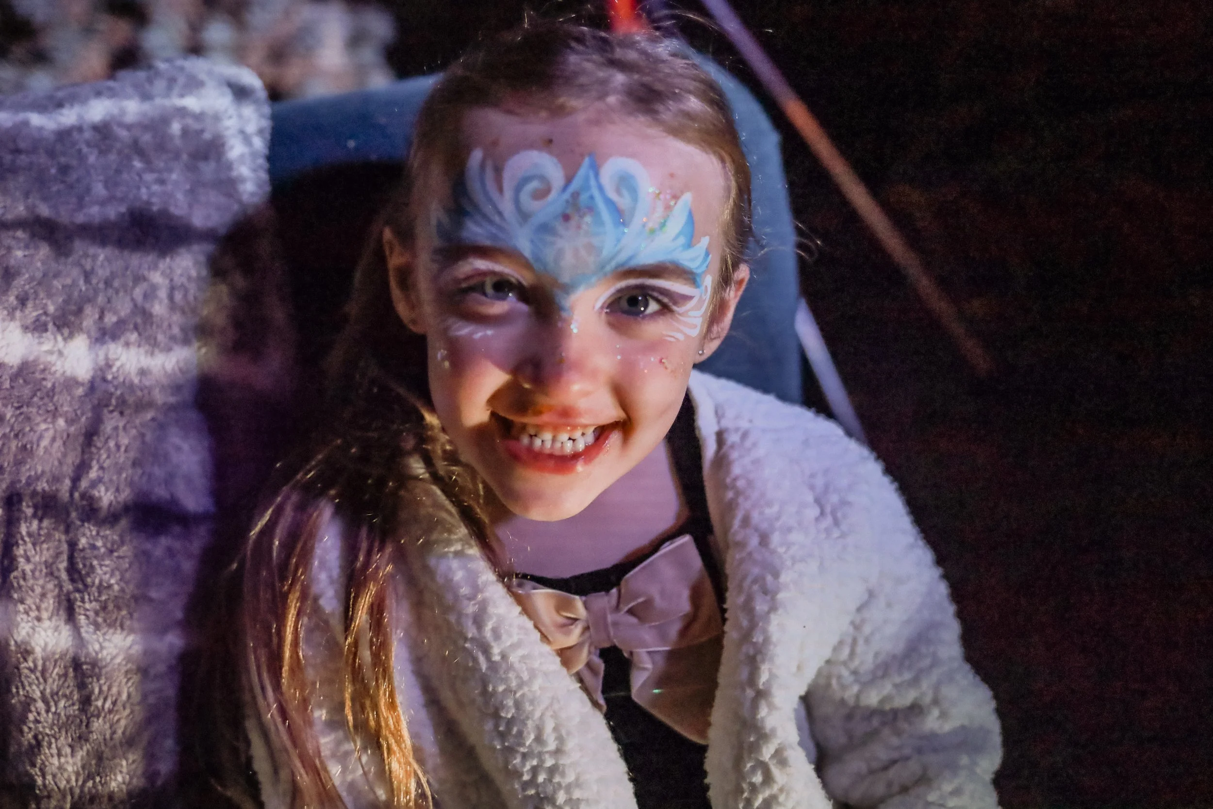A young girl with face paint in blue and white, glitter, and decorative designs, smiling. She has long brown hair and is wearing a white, fluffy jacket and a pink top with a bow. The background is dark, with some visible stone and wooden elements.