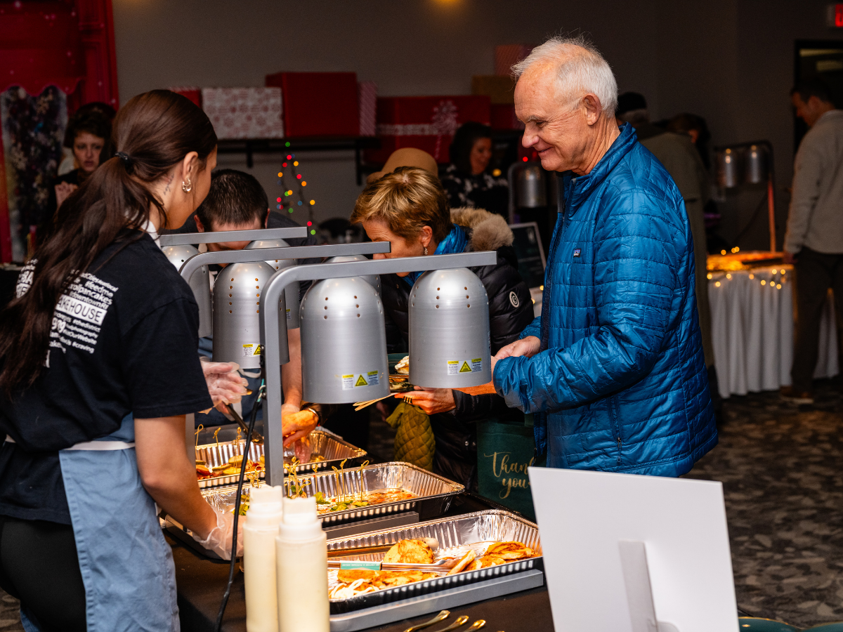 People serving and selecting food at a buffet line during a holiday event, with festive decorations and Christmas gifts in the background.