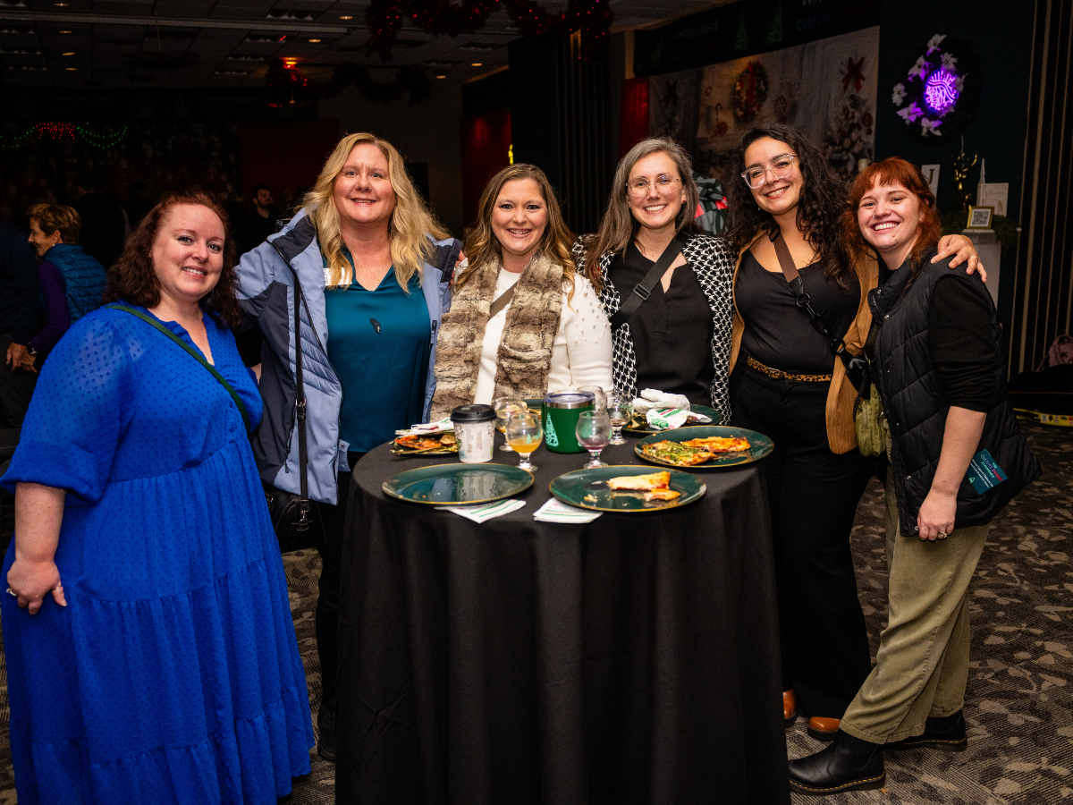 Six women standing around a table with some pizza and drinks, smiling at the camera, in a decorated indoor setting.