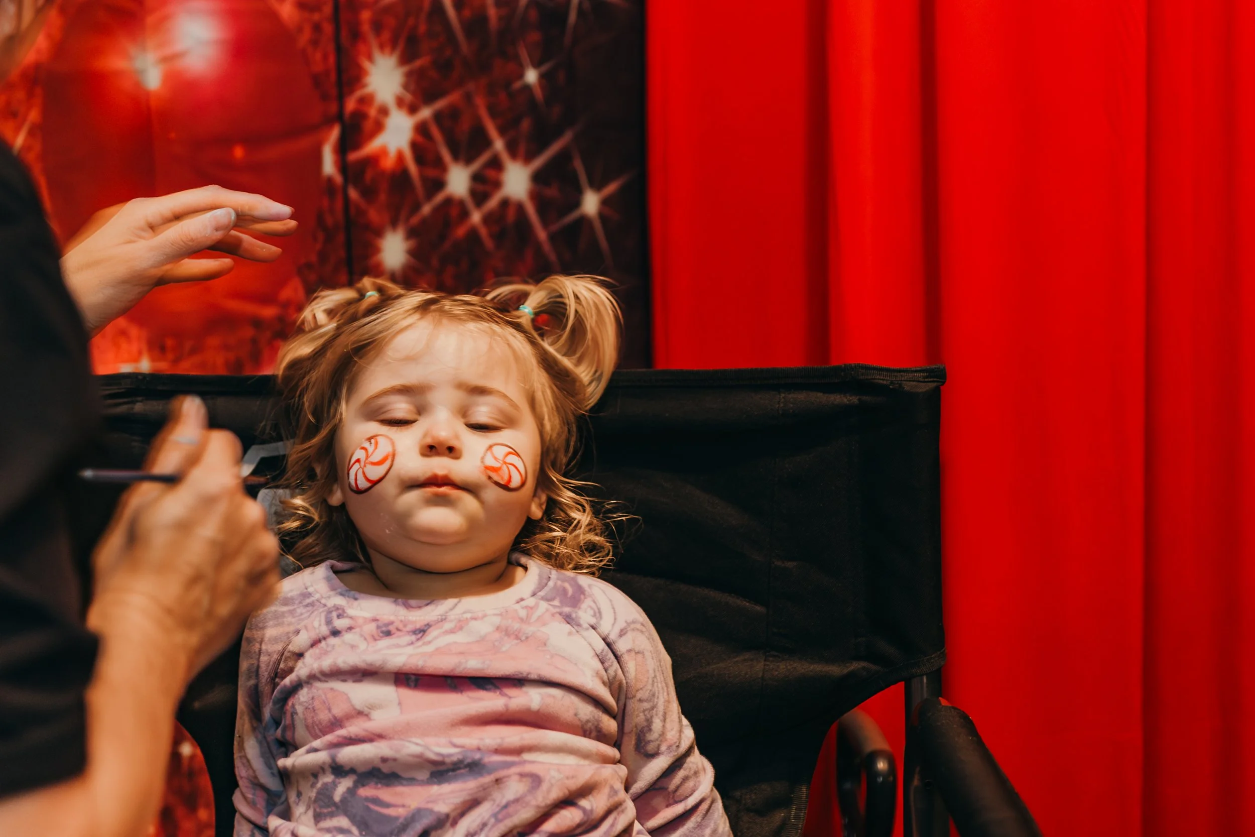 A young girl with face paint of red and white peppermint candies is having her face painted by an artist in a setting with red curtains and holiday-themed decorations.