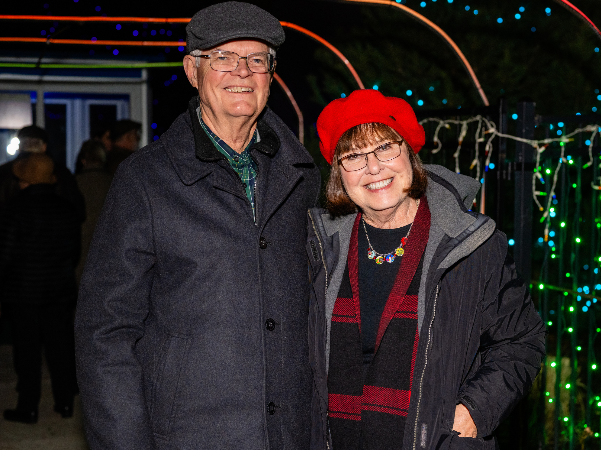 A smiling older man and woman posing together outdoors at night with colorful holiday lights in the background. The man wears glasses, a gray flat cap, and a dark coat. The woman wears glasses, a red beret, and a dark coat with a black and red scarf.