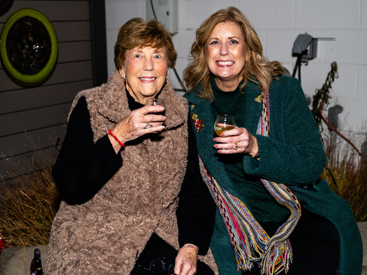 Two women sitting outside, smiling, and holding drinks. The woman on the left has short curly hair and is wearing a beige faux fur vest. The woman on the right has long wavy hair and is wearing a green cardigan with a colorful scarf.