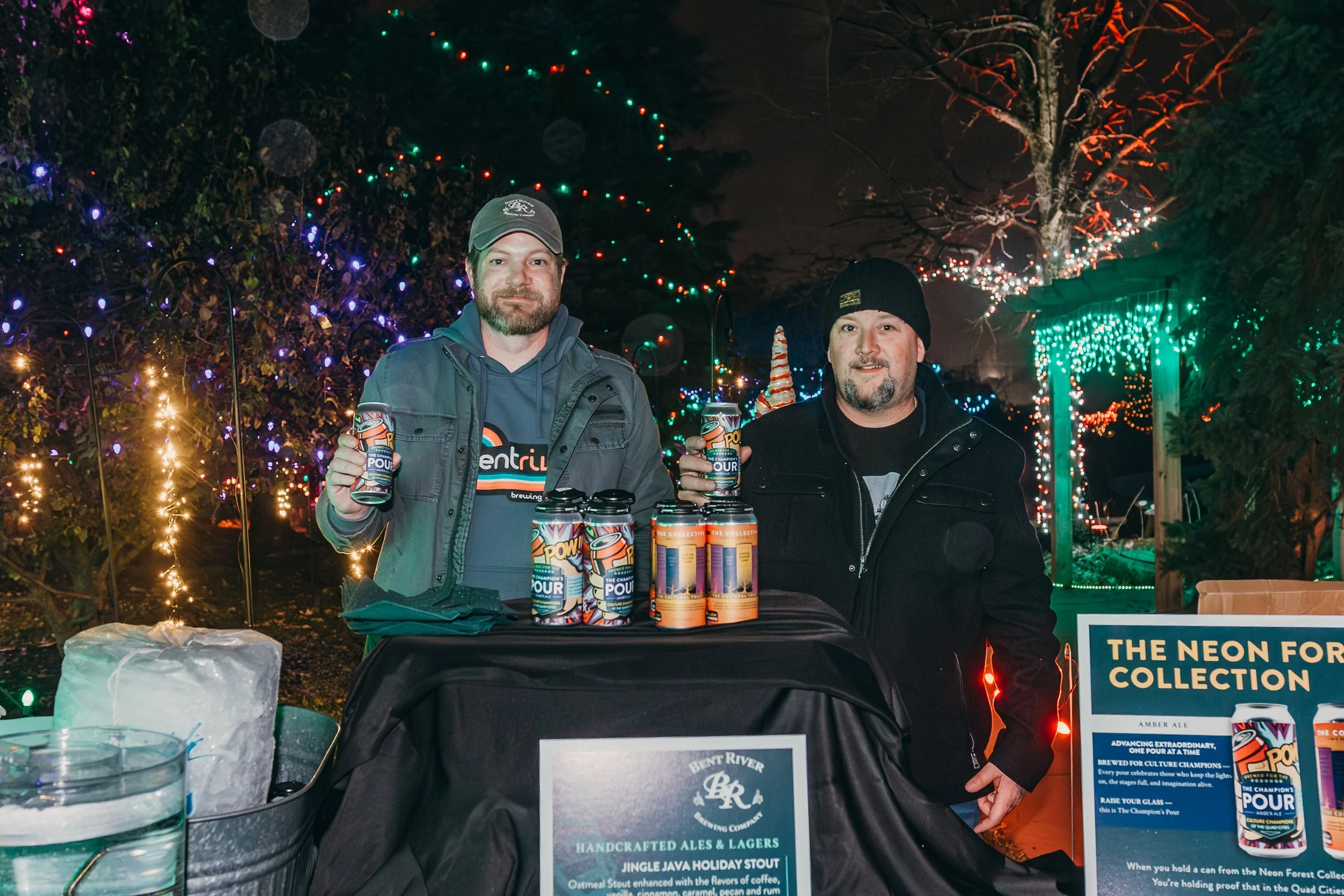 Two men standing behind a table with craft beers at night, decorated with colorful holiday lights and trees.