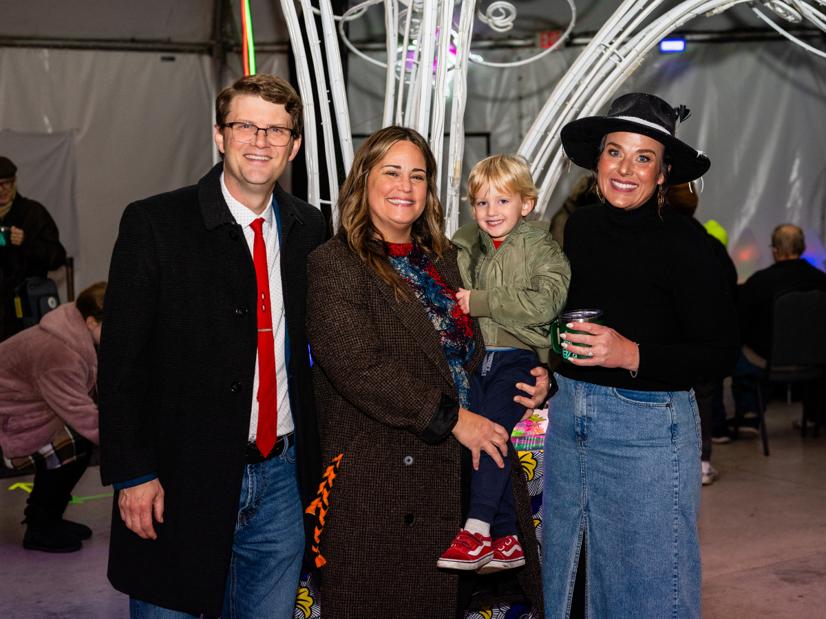 A group of four people, including a child, posing together indoors at an event or gathering, smiling at the camera. The group consists of a man in a dark coat and red tie, a woman with long hair in a brown coat, a young boy in a green jacket, and a w