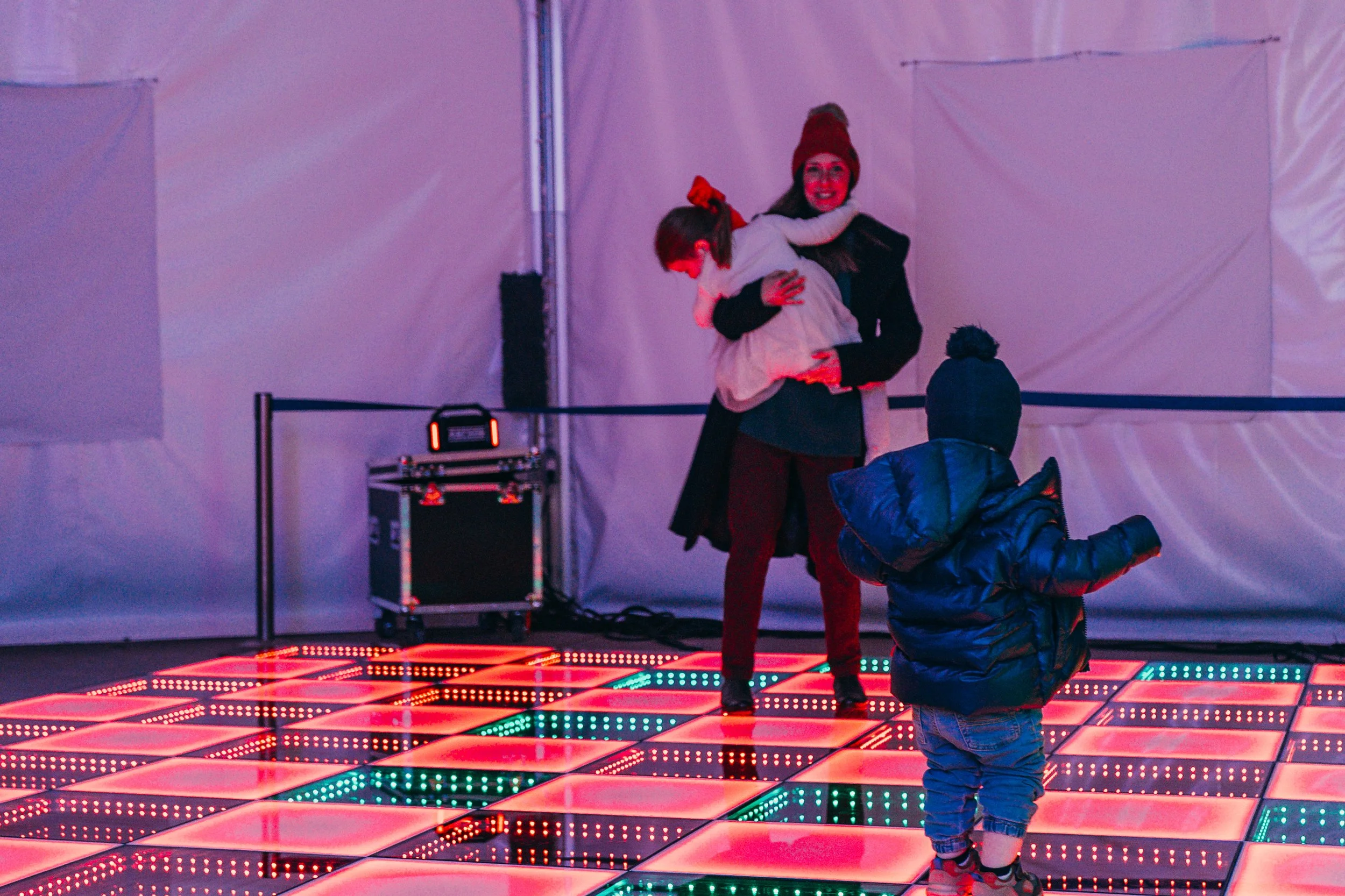 A woman holding a child in her arms on a colorful LED dance floor, with a young boy standing nearby. The woman is smiling and wearing a red beanie, while the child has a red bow in her hair and is hugging the woman. The boy is dressed in a blue jacke