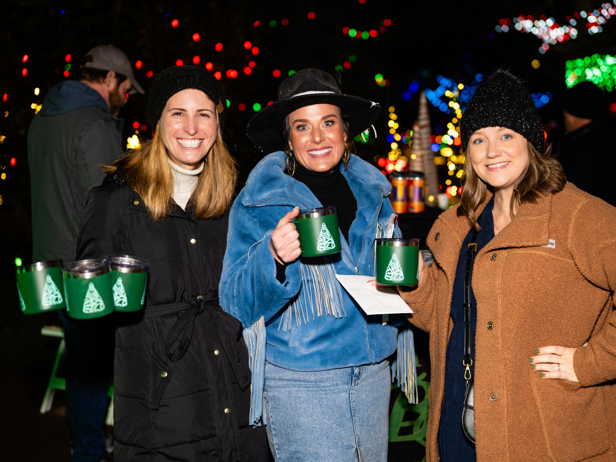 Three women smiling at a festive outdoor event during nighttime, holding mugs with Christmas tree designs, with colorful Christmas lights in the background.