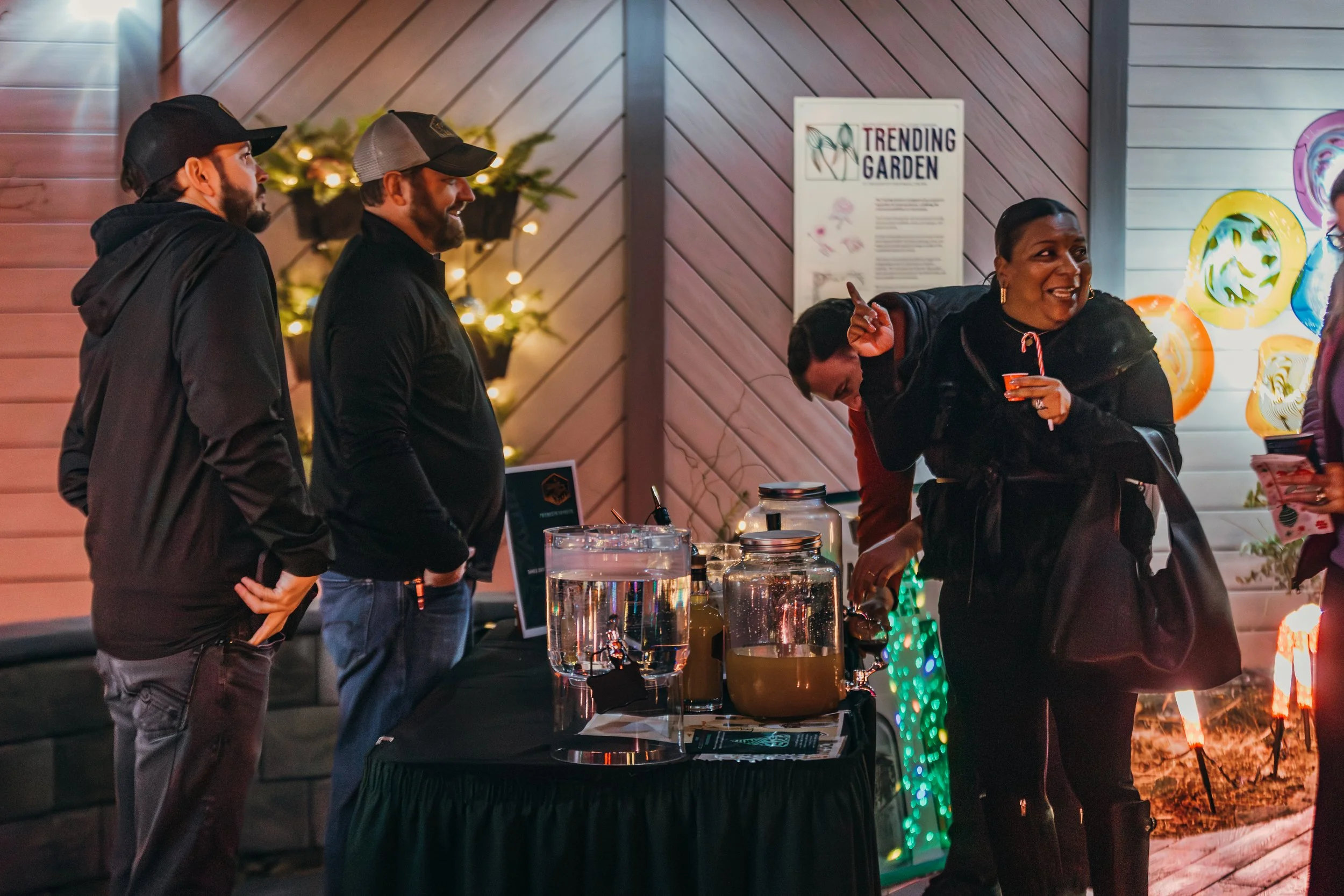 People at a booth with large drink containers, conversing and smiling in a festive outdoor setting with decorative lights and wall art.