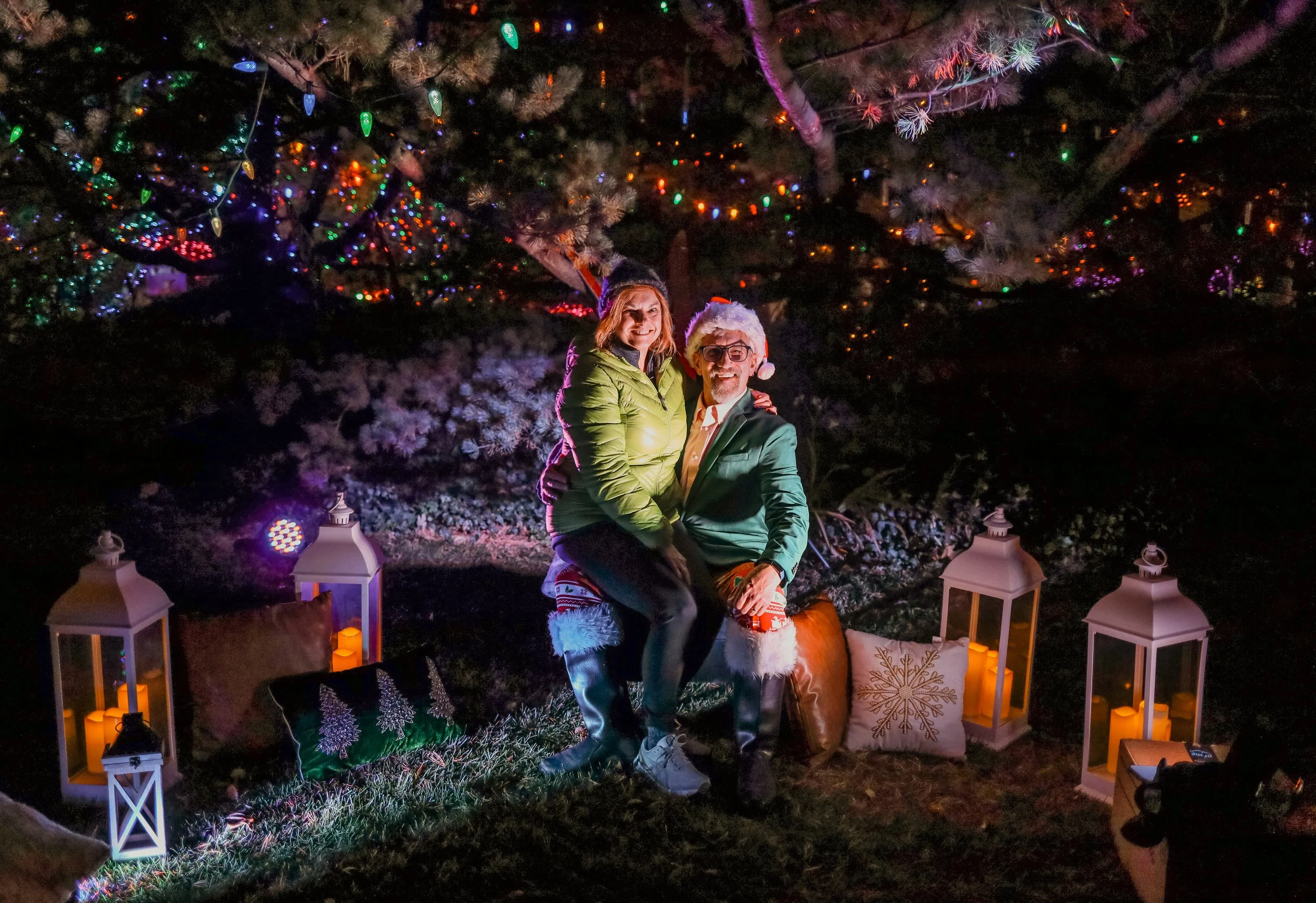 A couple dressed in Christmas-themed clothing, including a Santa hat and elf hat, sitting on a wooden log in front of decorated trees with colorful lights. They are surrounded by pillows, lanterns, and candles, creating a festive holiday outdoor scen