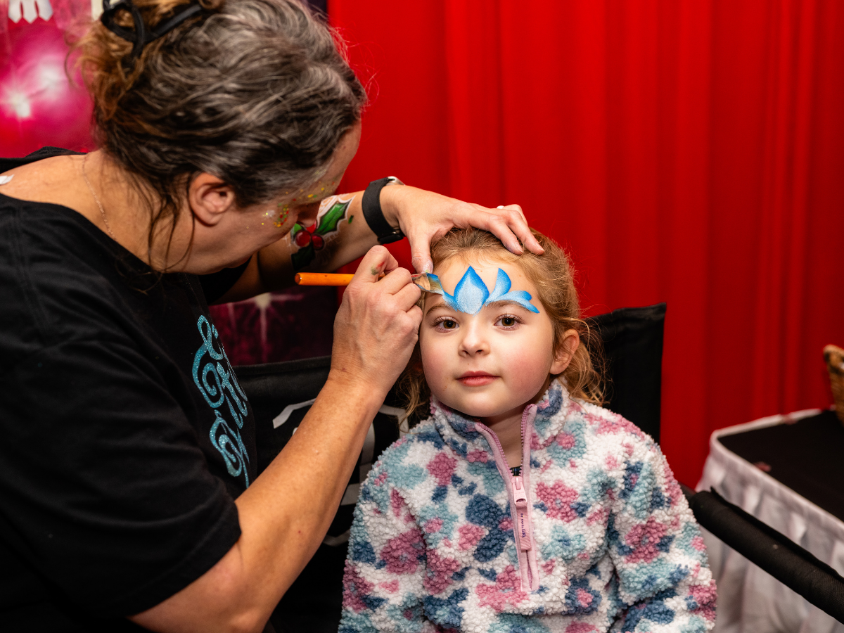 Young girl with face paint on her forehead, being painted by a woman. The face paint design is blue and resembles a flower or butterfly. The girl wears a colorful, patterned fleece jacket, and the woman has floral designs on her arm, with a red curta