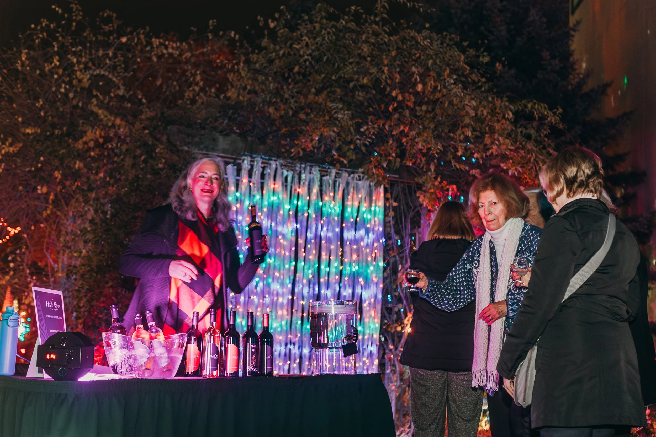 A woman with gray hair, wearing a black jacket and a colorful shawl, pours wine at an outdoor event at night with a backdrop of blue and multicolored string lights. Several people stand nearby holding glasses of wine, with trees and festive lighting 