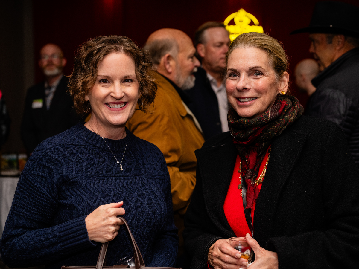 Two women smiling at a social event, with several people engaged in conversation in the background, indoors with warm lighting.