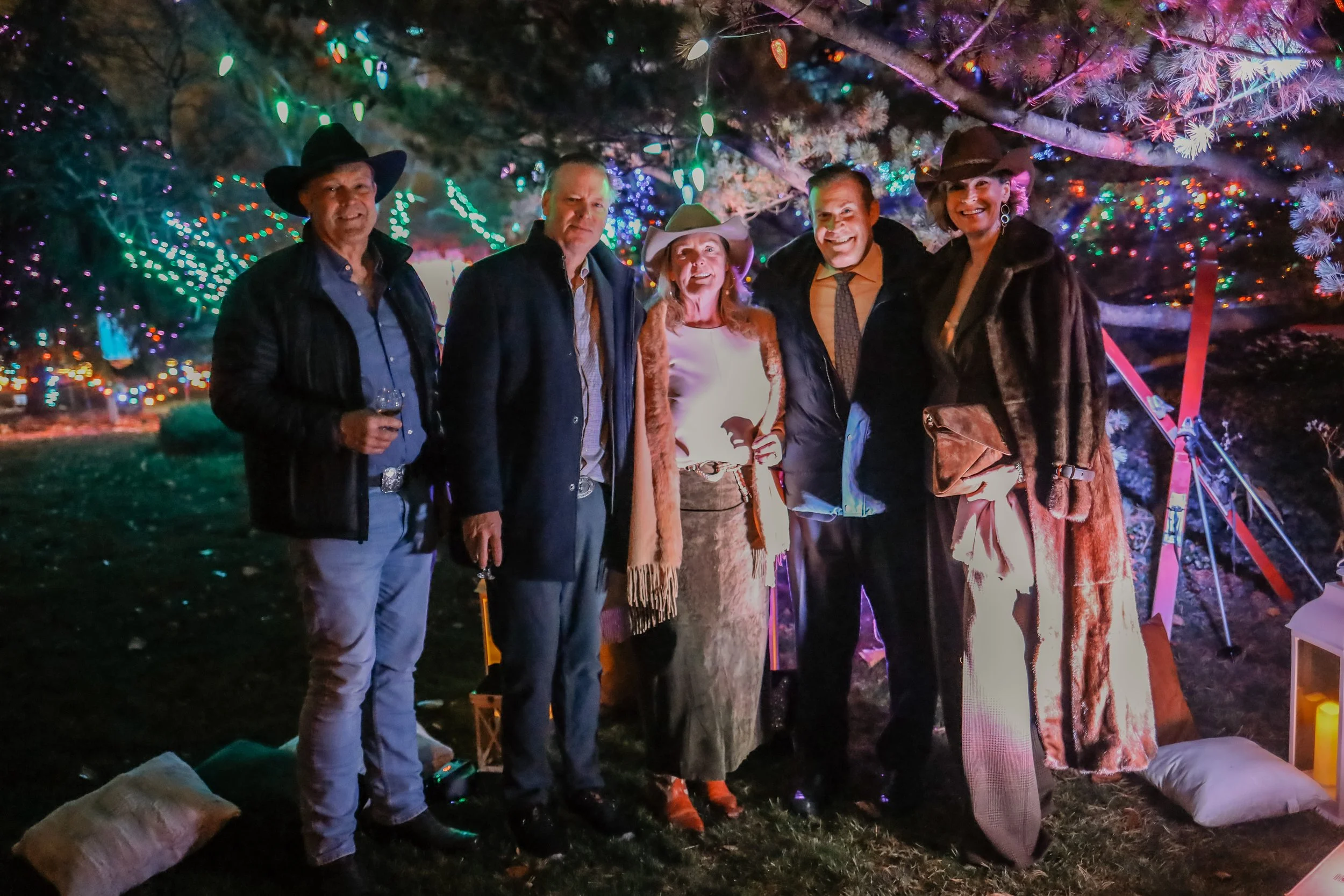 Group of six people dressed in formal and cowboy attire standing outdoors at night under a tree decorated with colorful string lights, with pillows and lanterns on the ground.