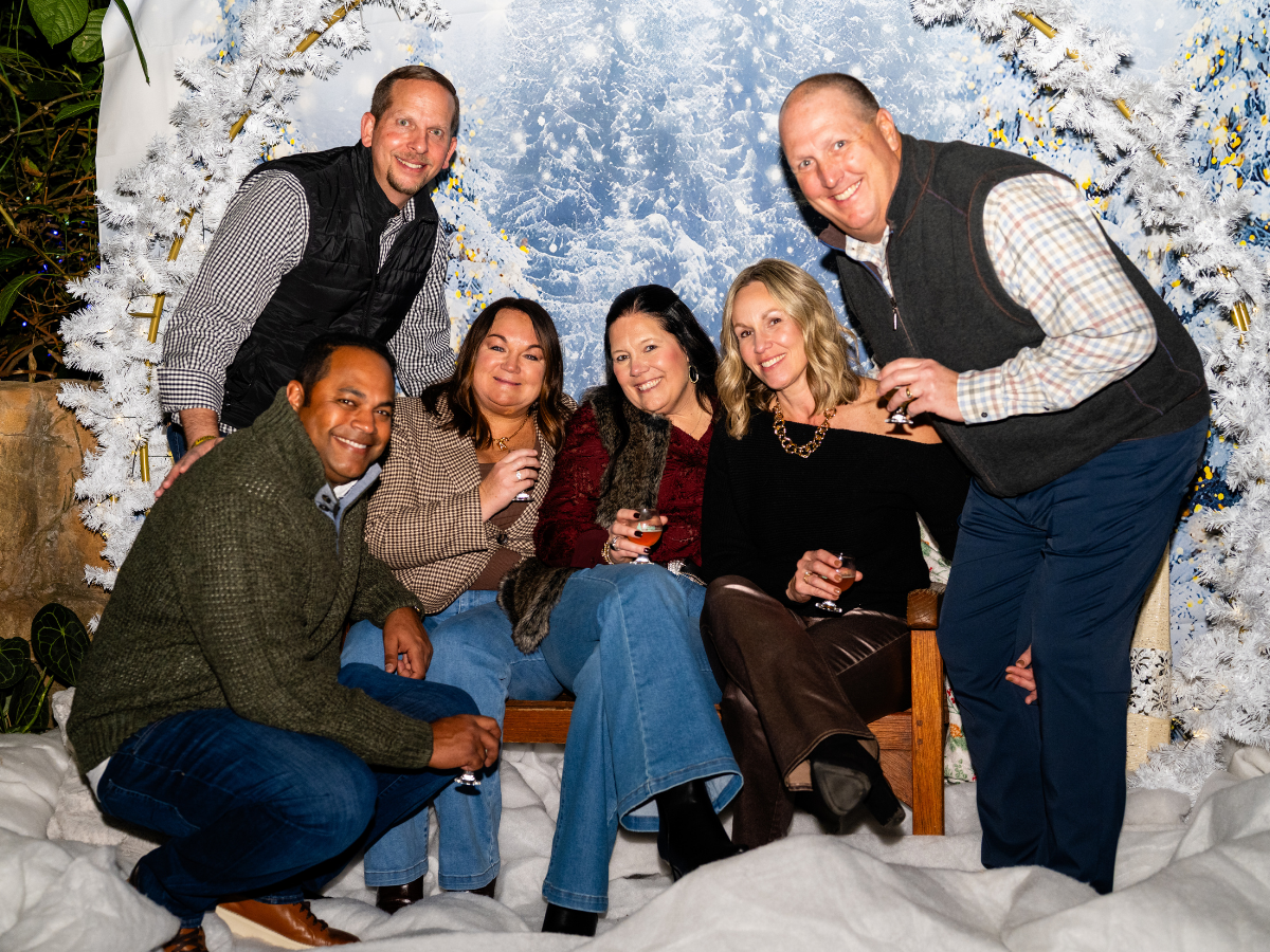 A group of six adults posing for a photo in front of a winter-themed backdrop with white trees and snow. They are smiling and holding drinks, appearing to enjoy a holiday celebration.