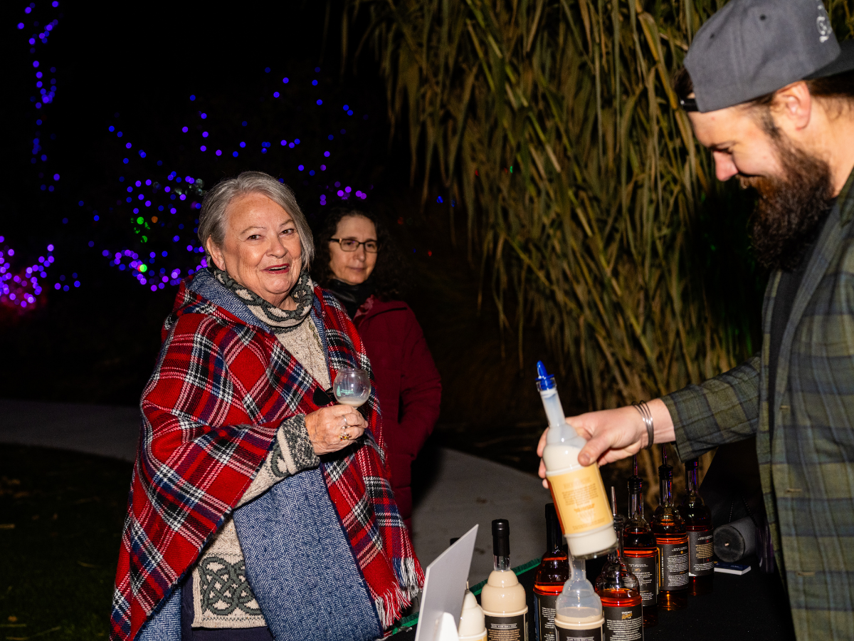 An elderly woman in a red plaid shawl holding a glass of wine at an outdoor event at night, with a man serving drinks and another woman in the background, surrounded by colorful decorative lights and foliage.