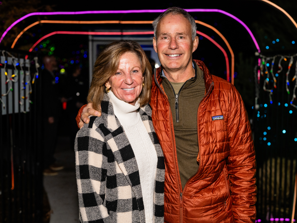 A smiling middle-aged woman in a black and white plaid coat and a cream turtleneck, standing next to a smiling middle-aged man in a red puffer jacket and olive green shirt, at night under colorful string lights.
