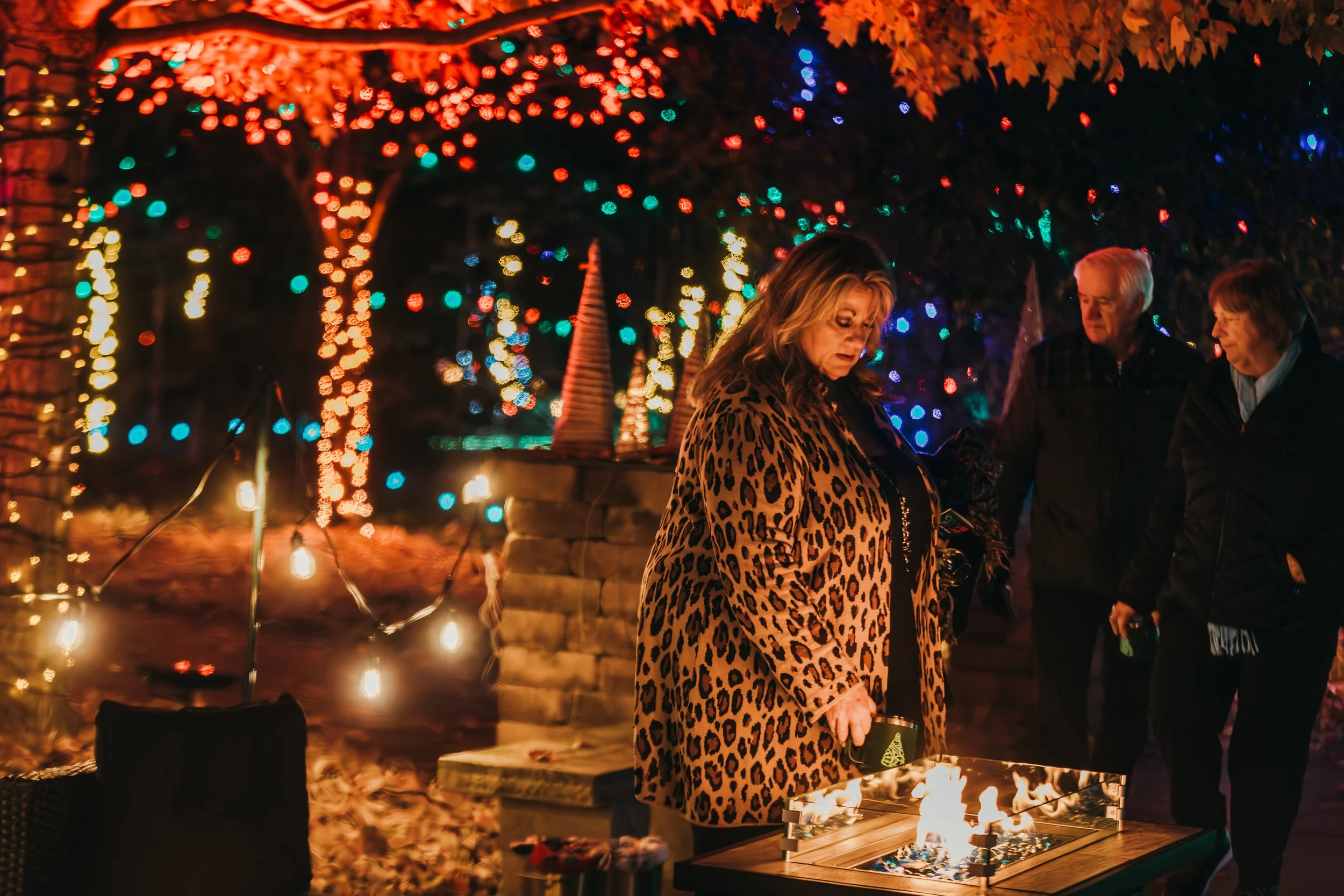 People gathered around a fire pit outdoors at night, decorated with colorful string lights and illuminated trees, enjoying a warm fire on a festive evening.