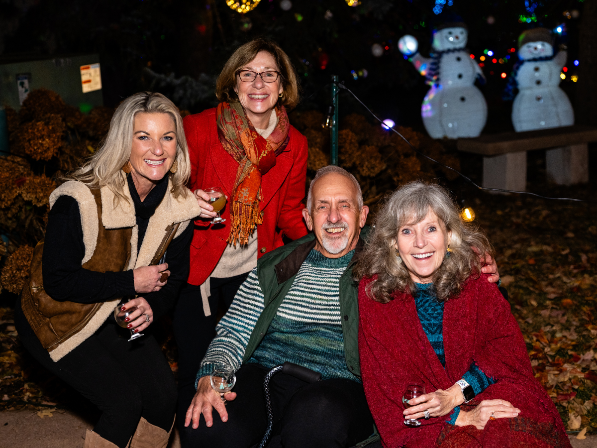 A group of five happy, smiling people at a festive outdoor gathering during Christmas, with Christmas lights and inflatable snowmen in the background, holding drinks.