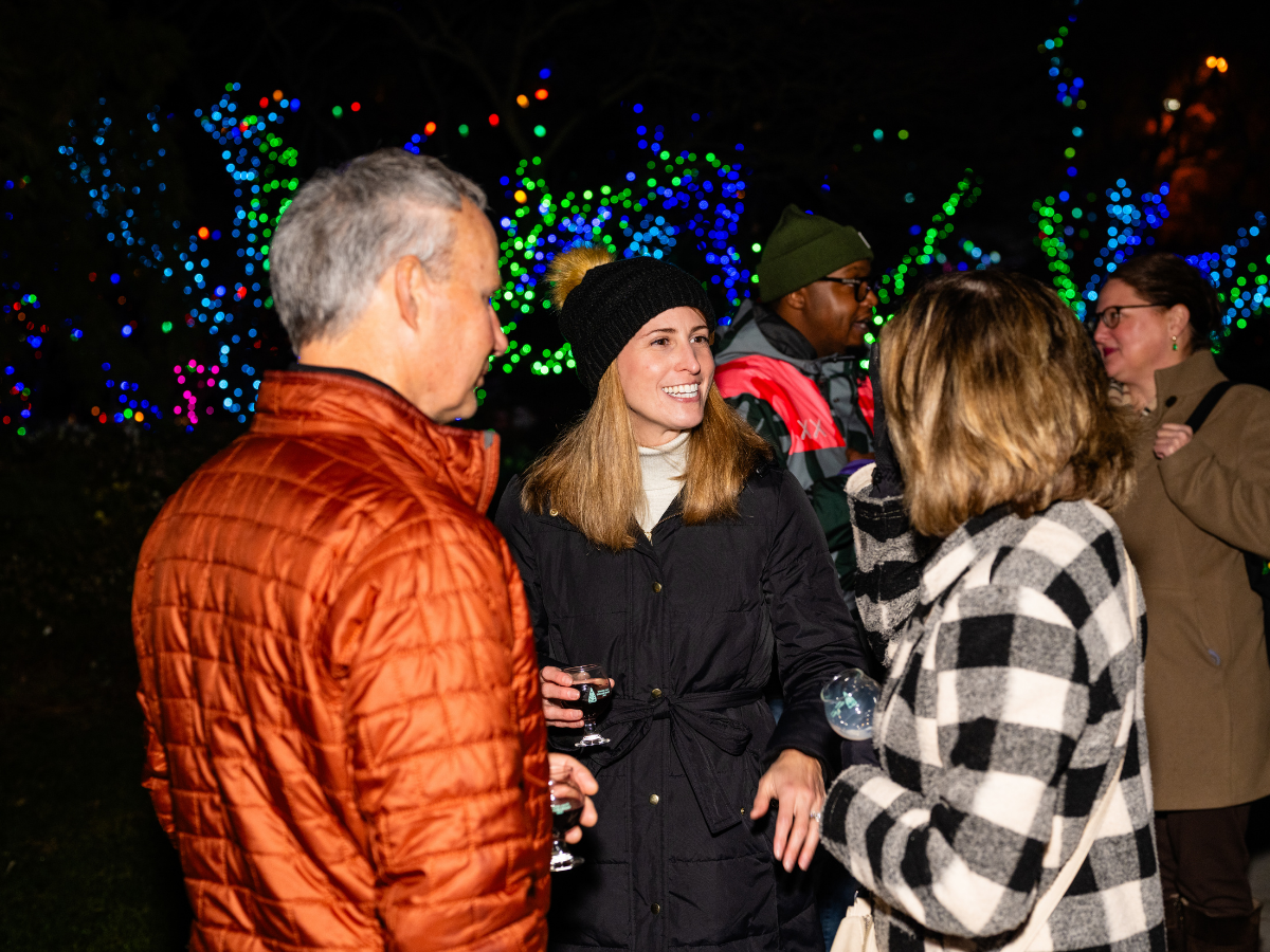 Group of adults outdoors at night, conversing, with colorful holiday lights in the background, some holding glasses.
