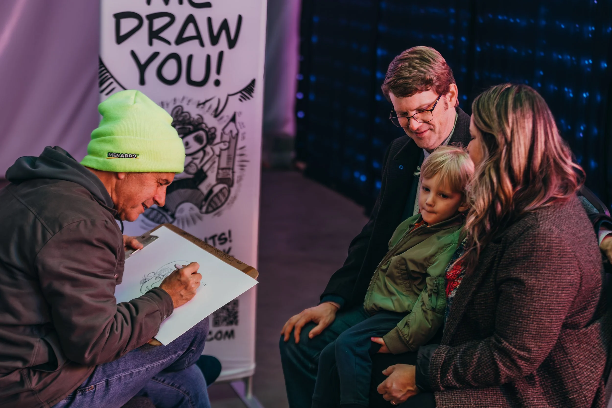 A man with a yellow beanie and dark jacket draws a caricature of a family sitting on a bench at an event or convention.