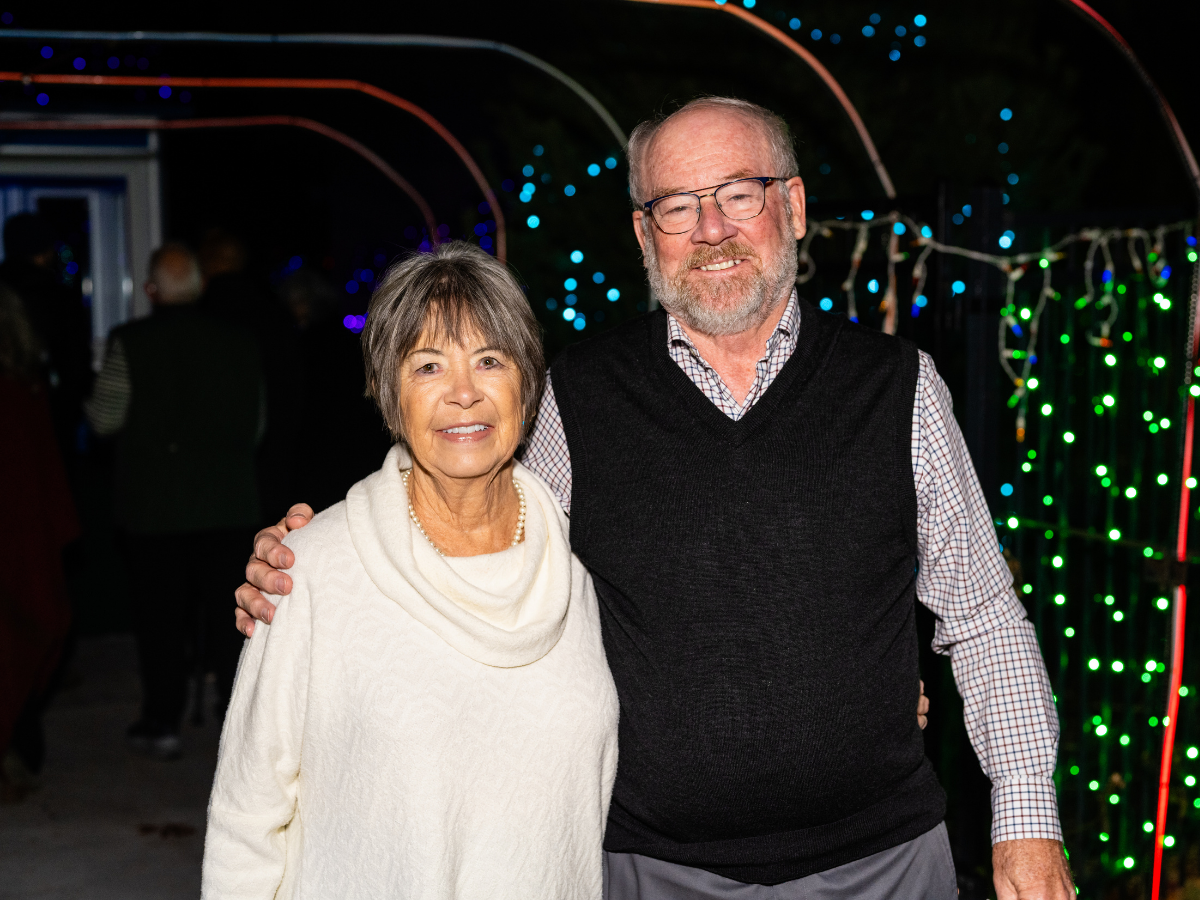 A smiling older woman and man standing close together at an outdoor event at night, surrounded by colorful string lights.
