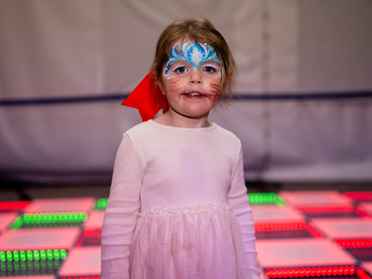 Young girl with face paint resembling a snowflake and cat whiskers, wearing a pink dress with a red bow in her hair, standing in front of a colorful, illuminated background.