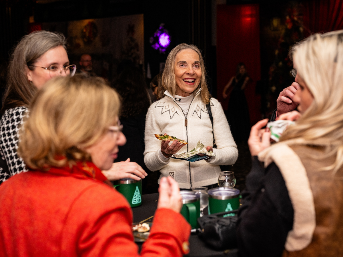 Group of women smiling and talking at a social gathering, with food and drinks on the table.