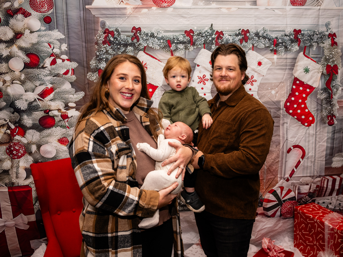 Family of four celebrating Christmas indoors with decorated tree, stockings, and presents, holding two young children.