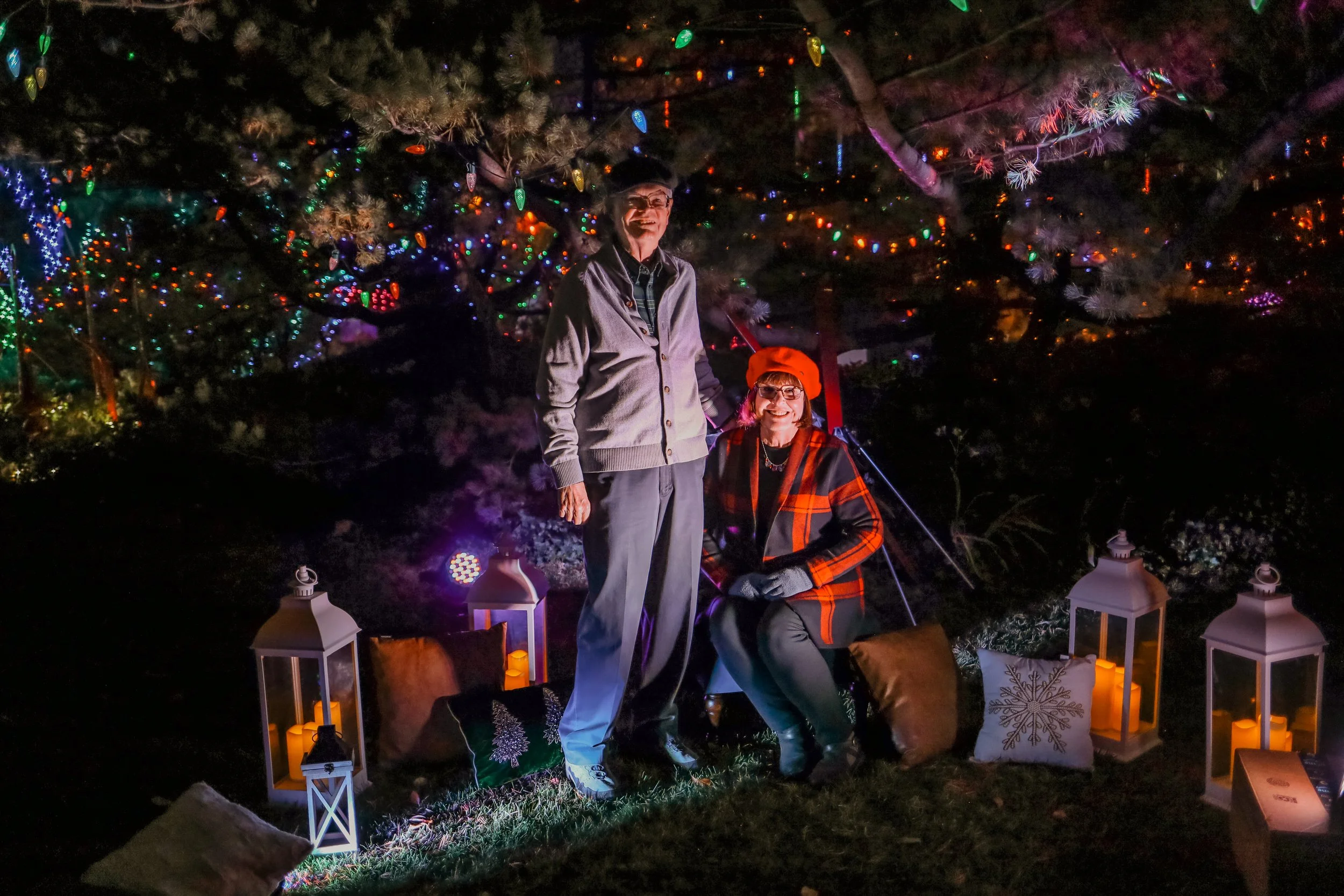 Two women in fall or winter clothing with hats and gloves, sitting and standing in a festive outdoor setting at night, surrounded by Christmas decorations, including lanterns with candles, pillows, and colorful string lights on trees.