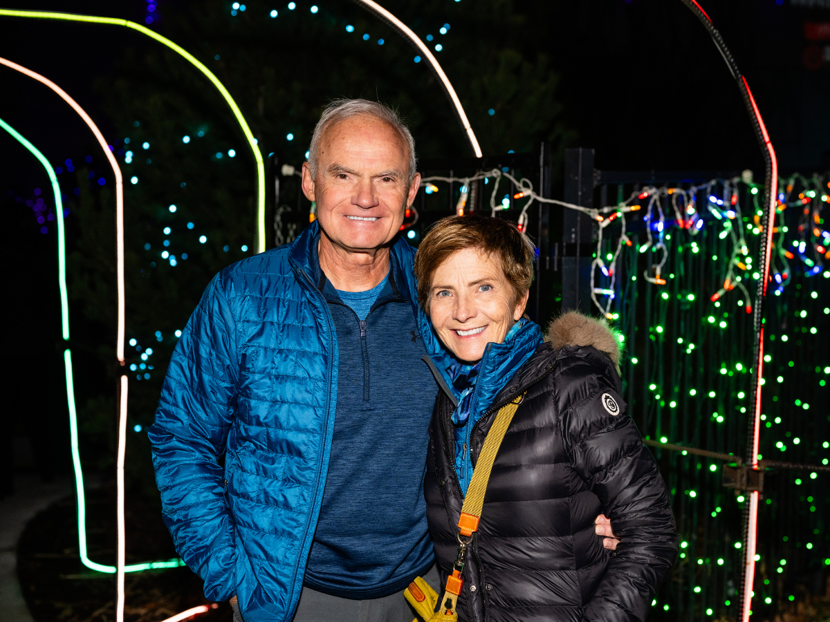 An older man and woman standing outside at night near colorful holiday lights, smiling at the camera, both wearing blue and black jackets.