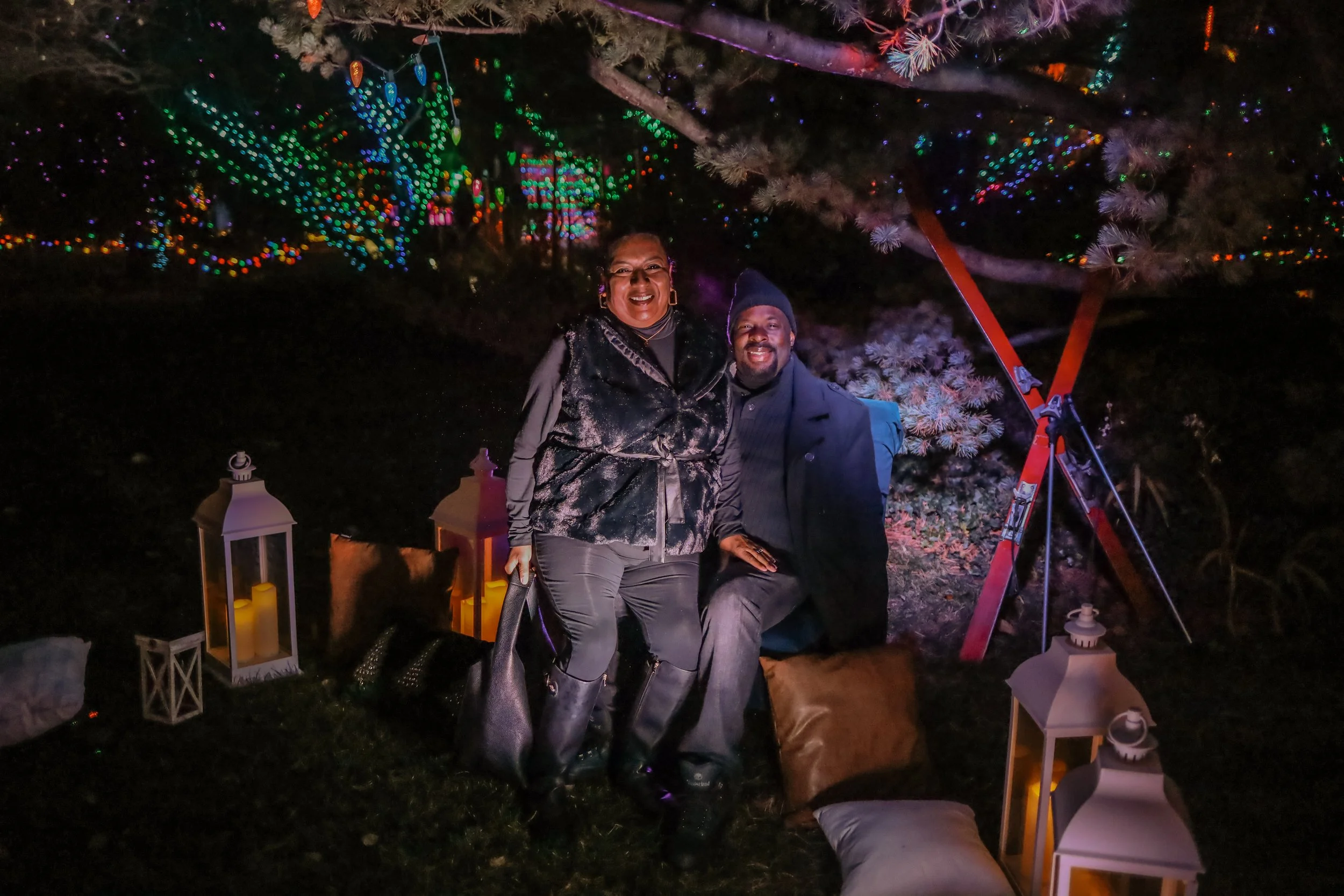Two people sitting on a stack of pillows and cushions outdoors at night, surrounded by decorated lanterns and colorful string lights, with a background of illuminated trees and a large telescope.