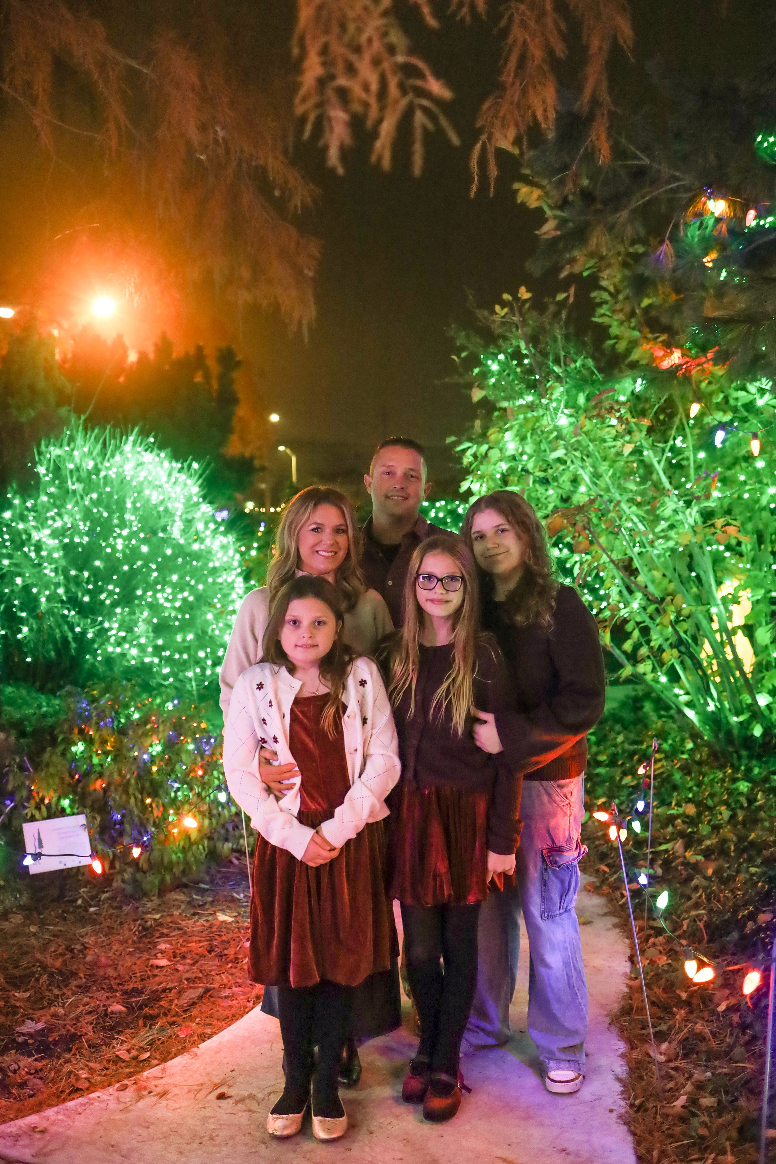 A family of five, including three girls and two adults, stands together on a pathway surrounded by colorful Christmas lights and greenery at night.
