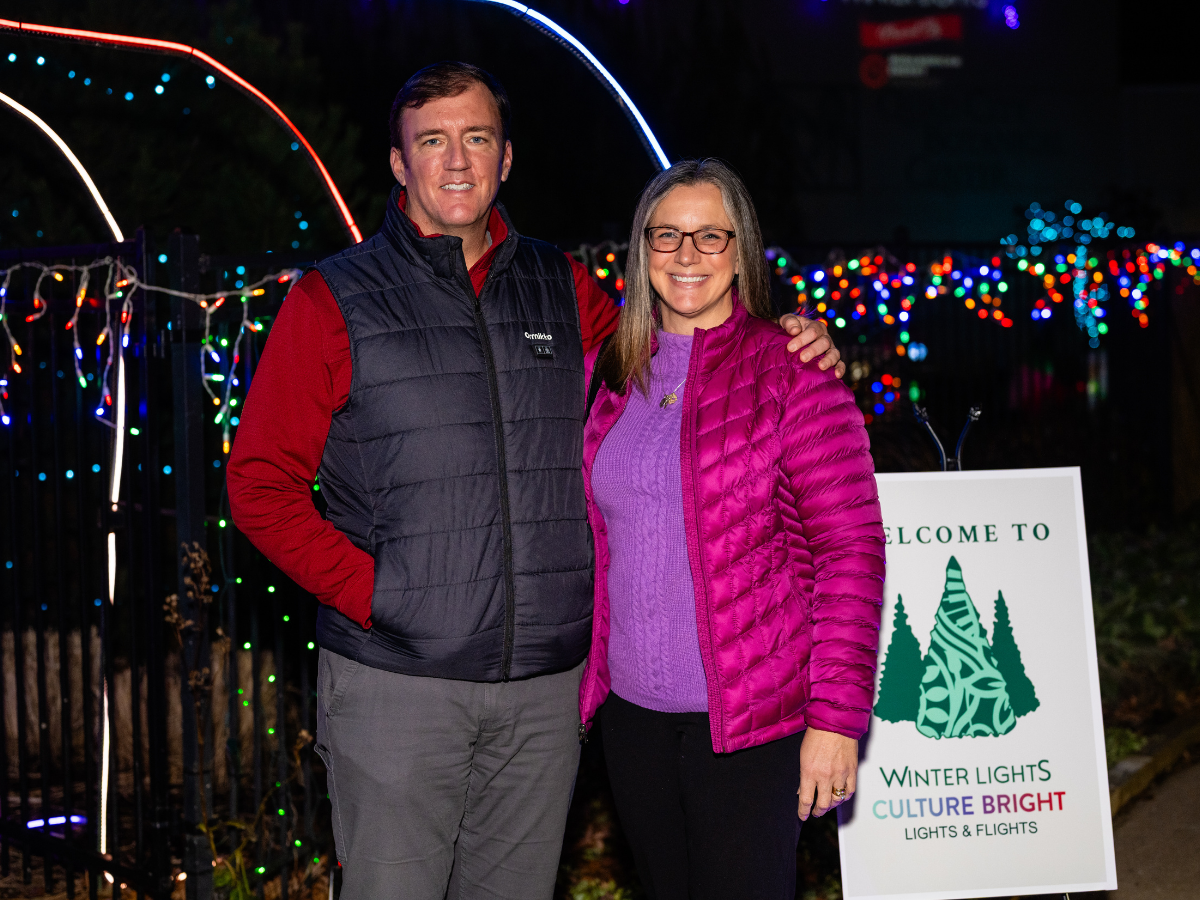 A man and woman smiling and posing outside at night with colorful holiday lights behind them. The woman is wearing glasses and a pink puffer jacket, and the man is wearing a black vest over a red long-sleeve shirt. They stand next to a sign that read