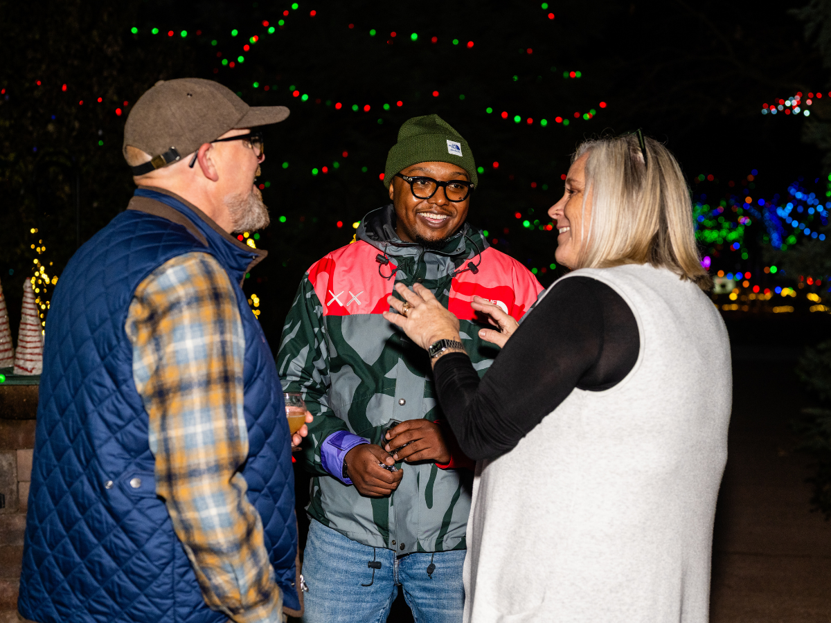 Three people talking outdoors at night with colorful Christmas lights in the background.