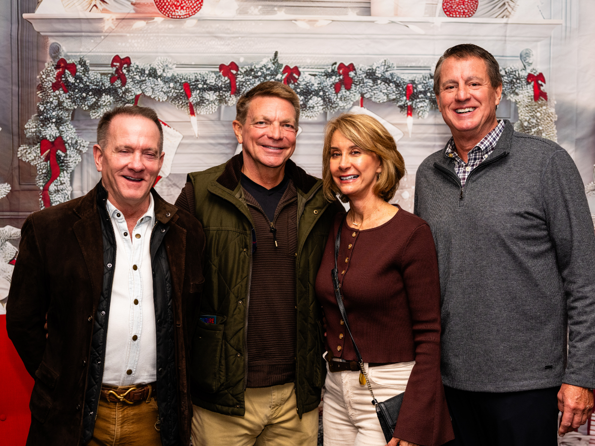 Four adults posing in front of a holiday-themed fireplace decorated with Christmas greenery, red ribbons, and stockings.