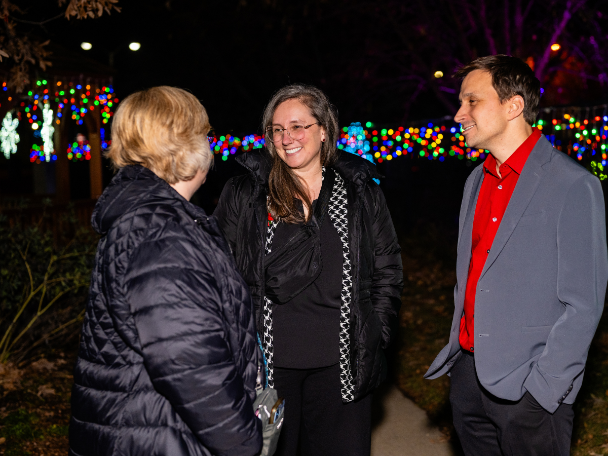 Three people having a conversation outdoors at night, with colorful Christmas lights in the background.