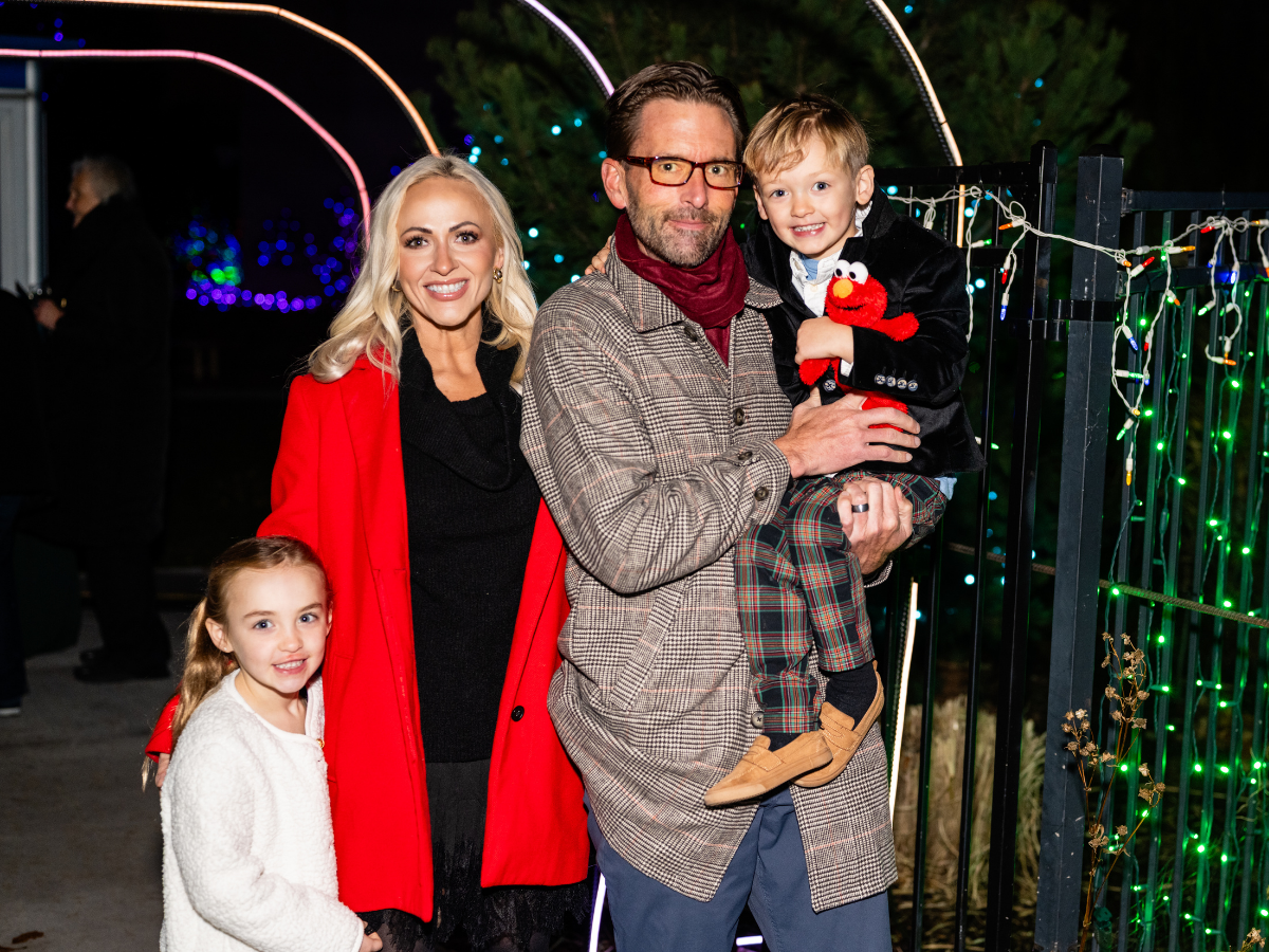 A family of four smiling at night near holiday Christmas lights. The woman wears a red coat, and the young girl wears a white sweater. The man holds a young boy wearing a black jacket and plaid pants, holding a plush Elmo toy.