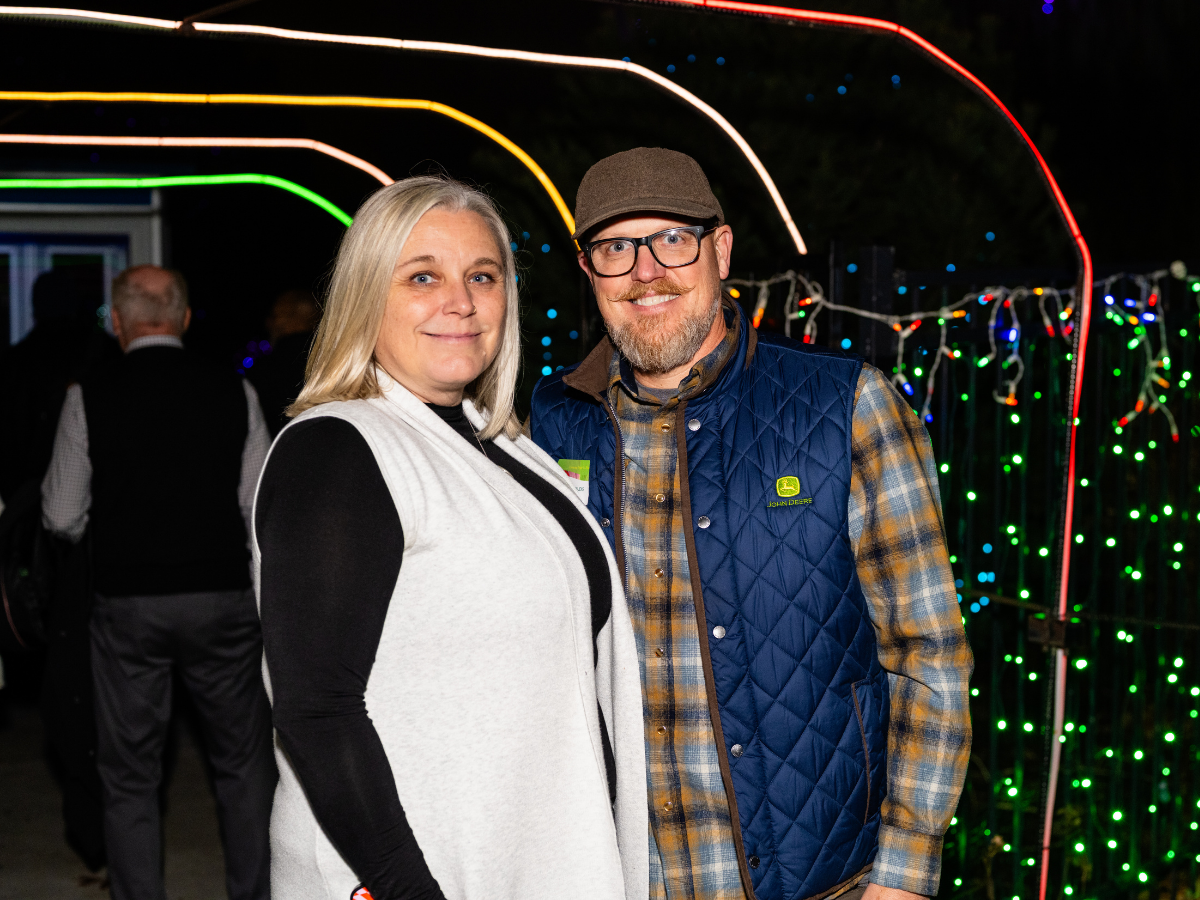 A man and woman standing outdoors at night, smiling, with colorful string lights in the background.