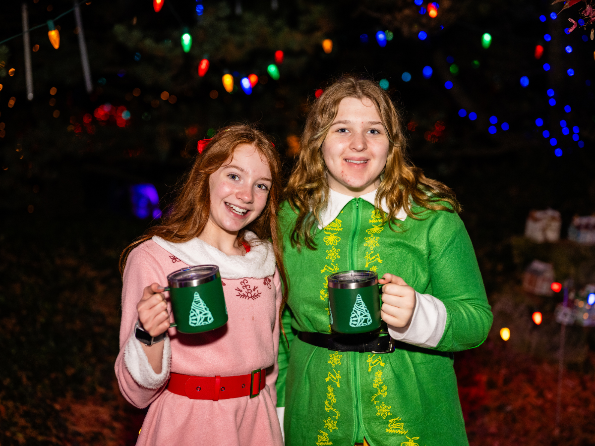 Two young girls in Christmas-themed outfits holding mugs with Christmas tree designs, standing in front of a decorated tree with colorful lights.