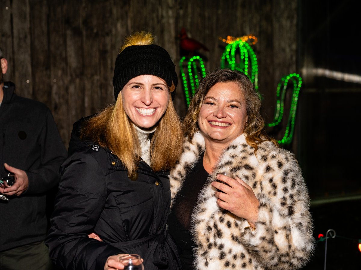 Two women smiling and posing for a photo at night, one wearing a black jacket and beanie, the other in a leopard print coat, with colorful cactus-shaped neon lights in the background.