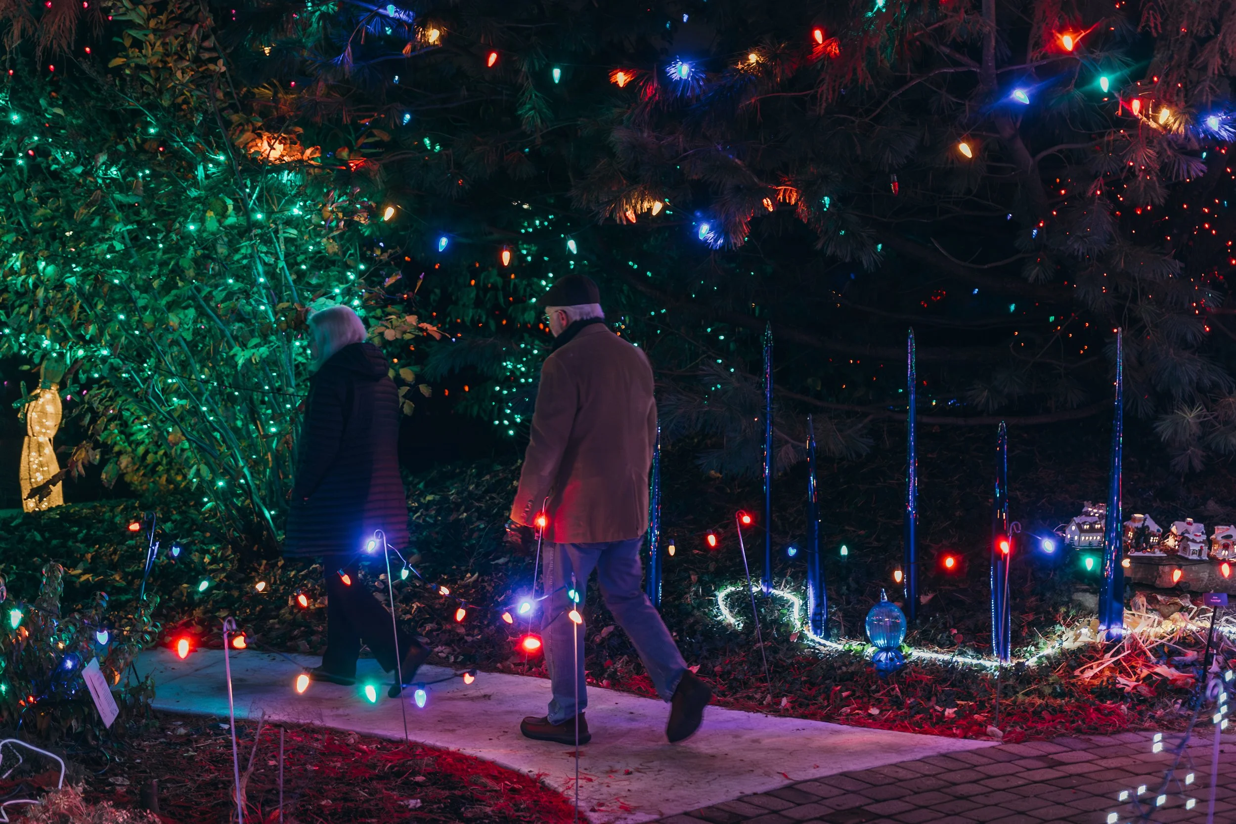 Two people walking on a decorated Christmas light display at night, with multicolored lights on trees and decorations around them.