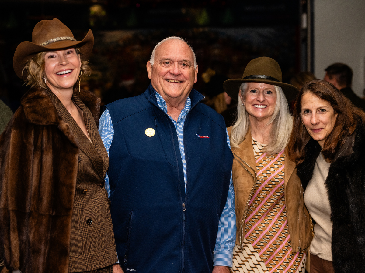Four people smiling at an event, dressed in casual and western-style clothing, with two women wearing hats and a man in a vest.