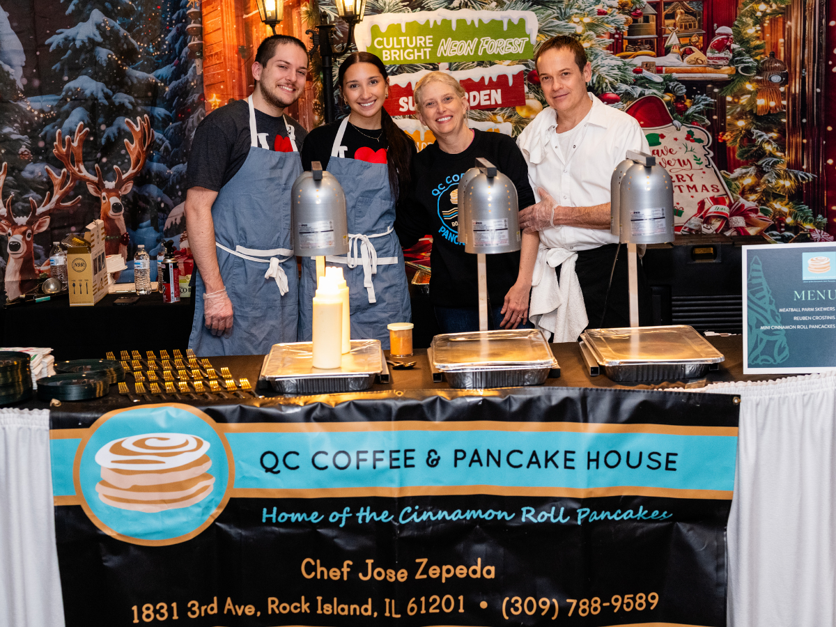 Four people standing behind a stall at a food booth for QC Coffee & Pancake House, with a holiday-themed background featuring reindeer and festive decorations. Two of them are wearing blue aprons, and the others are in casual clothing. The stall disp