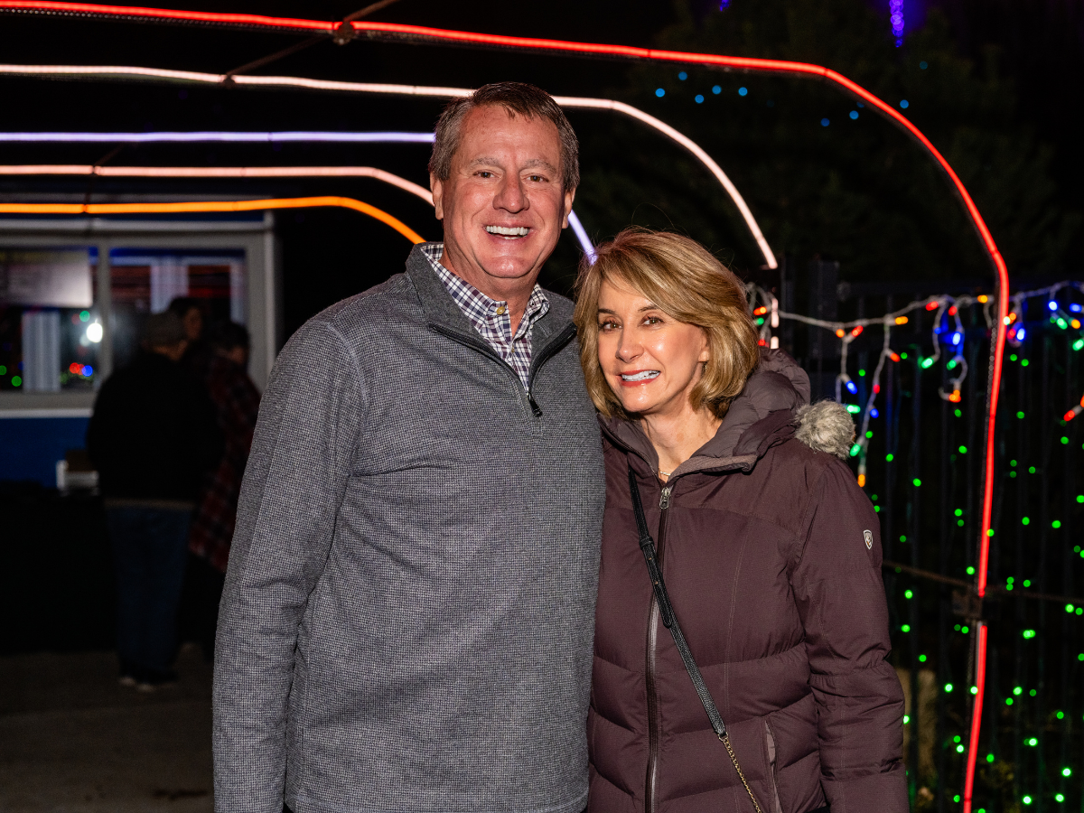 A smiling man and woman in warm jackets at a nighttime outdoor event with colorful string lights and neon lighting.