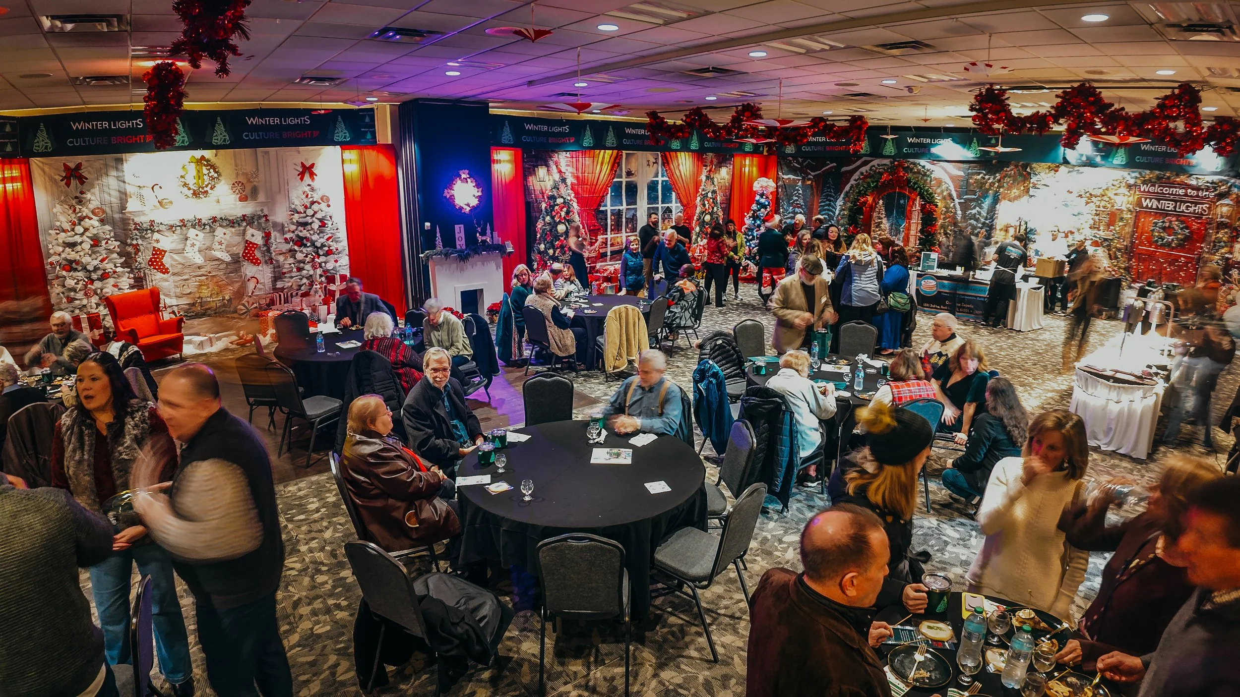 Indoor holiday event with decorated Christmas trees, festive wall backdrop, and people sitting and socializing at round tables, some eating and drinking. Red tinsel and holiday decorations adorn the ceiling.