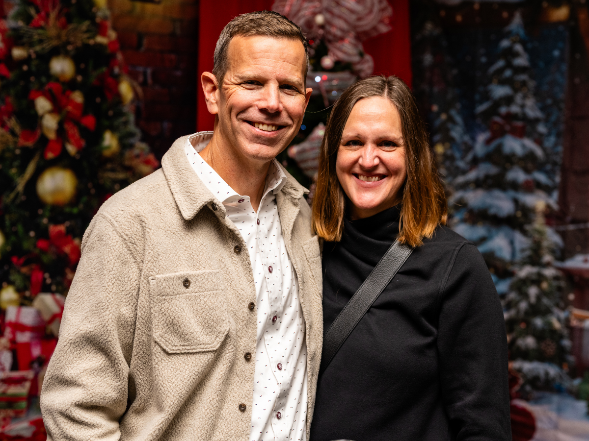 A man and woman smiling in front of decorated Christmas trees with ornaments and presents.