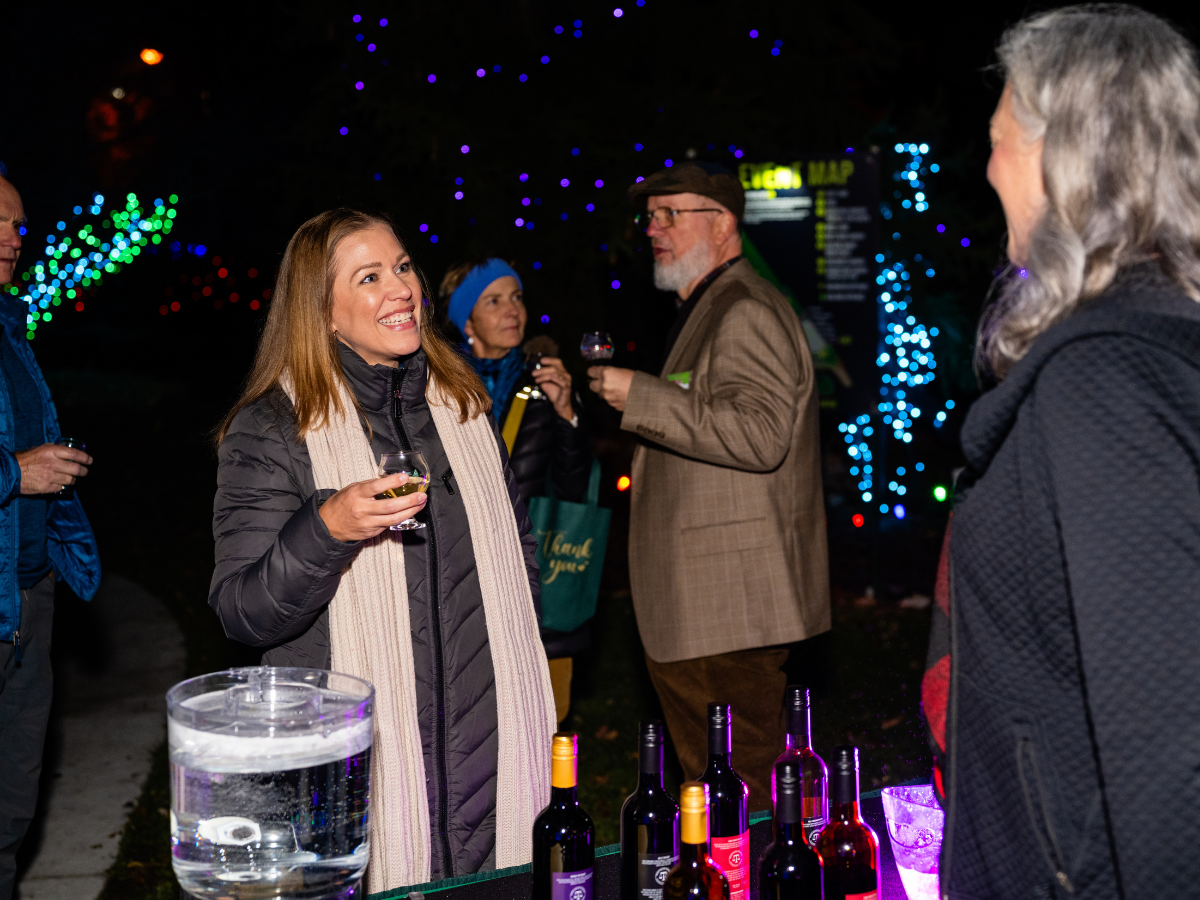 People socializing at an outdoor night event with string lights, holding drinks, near a table with wine bottles and a water pitcher.