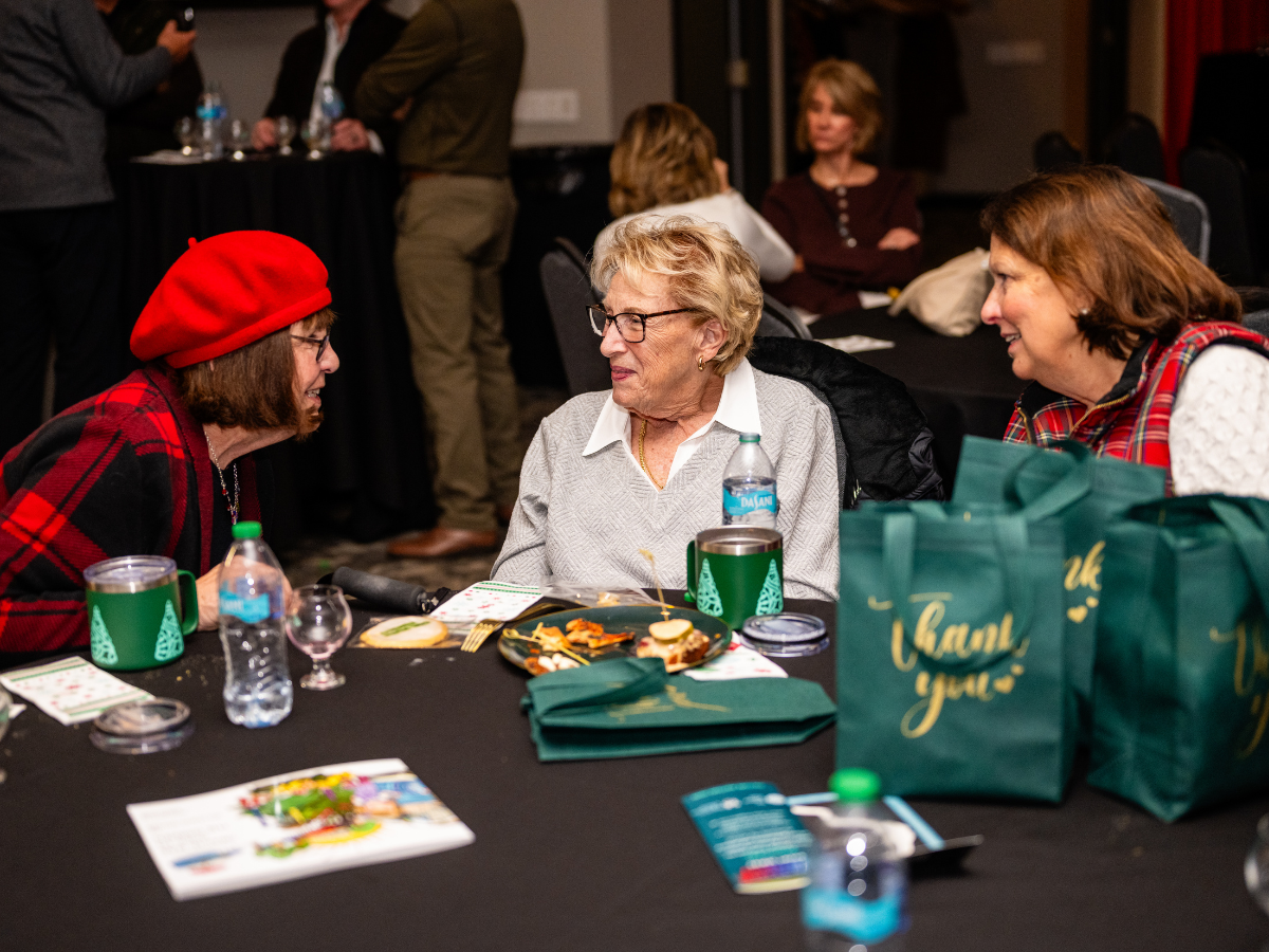 Three women sitting at a round table engaged in conversation, with holiday-themed gift bags and snacks on the table, in a festive indoor setting.