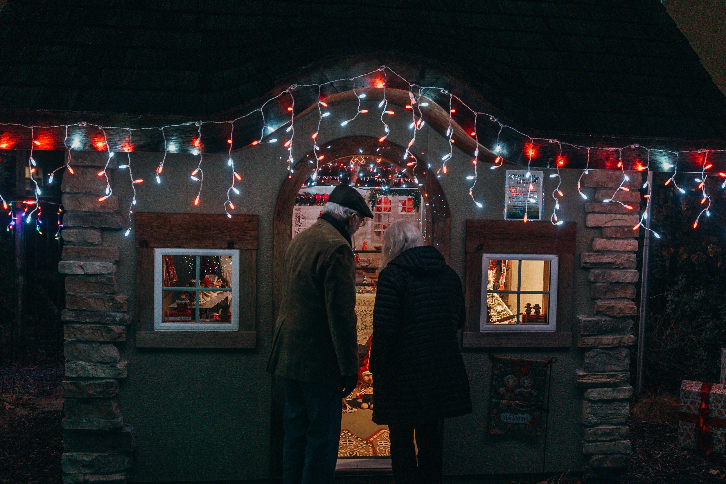 Two people, an elderly man and woman, stand close together outside a small house decorated with Christmas lights at night, talking or looking inside.