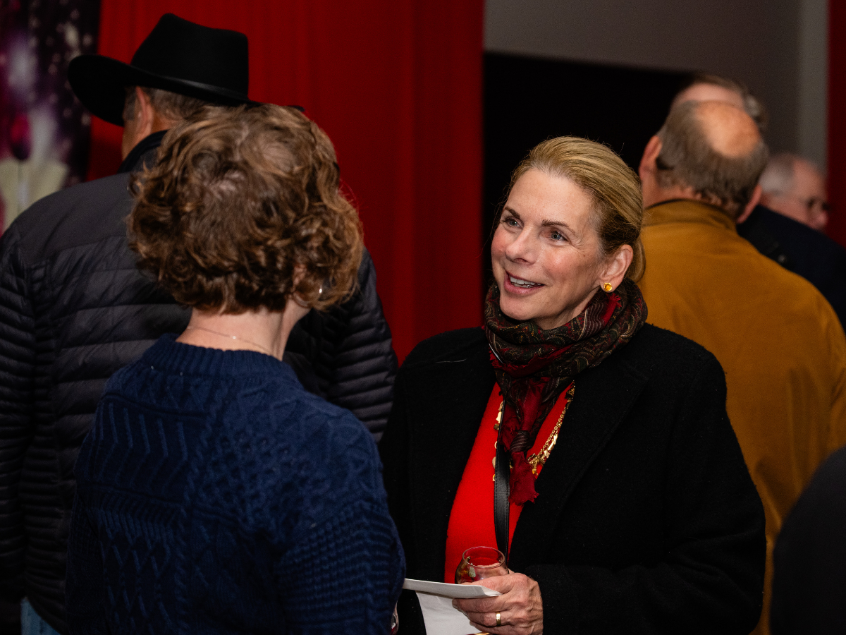 Two women are engaged in conversation at a social event. One woman has short, curly hair and is wearing a blue sweater, while the other woman has blonde hair in a bun, is dressed in a black coat with a red top, and is holding a glass in her hand. Oth