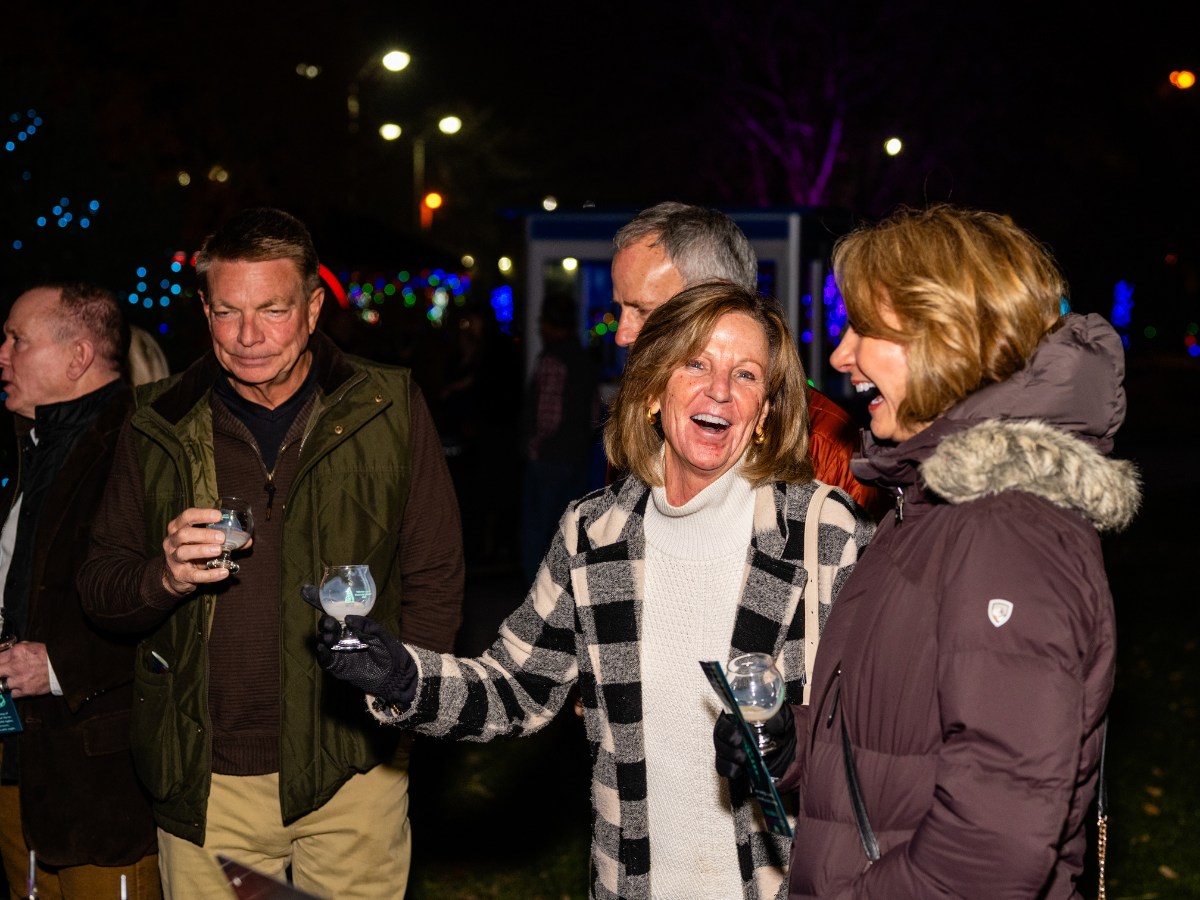 Group of people enjoying a social gathering outdoors at night, with colorful lights in the background, some holding drinks and engaging in conversation.